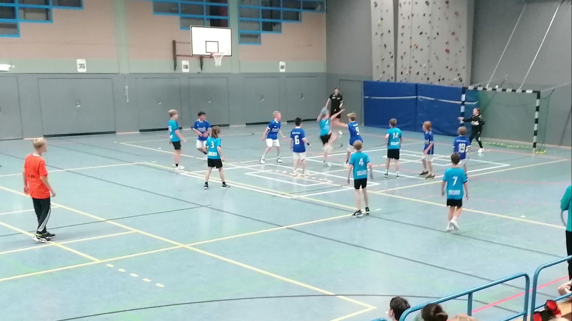 Jungen in blauen, türkisen und orangefarbenen Trikots spielen auf einem Hallenfeld Handball
