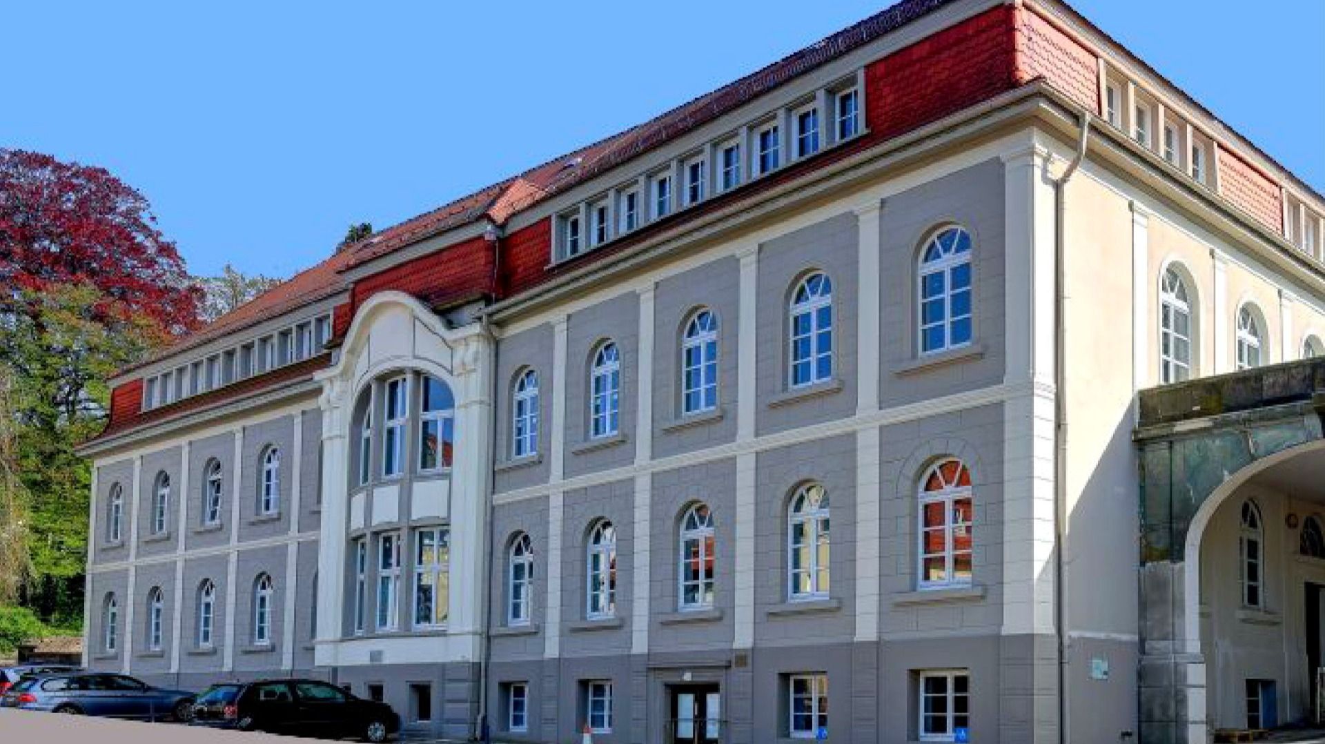 Mehrstöckiges Gebäude mit grauer Fassade, hellen Verzierungen, vielen Bogenfenstern, rotem Dach und parkenden Autos