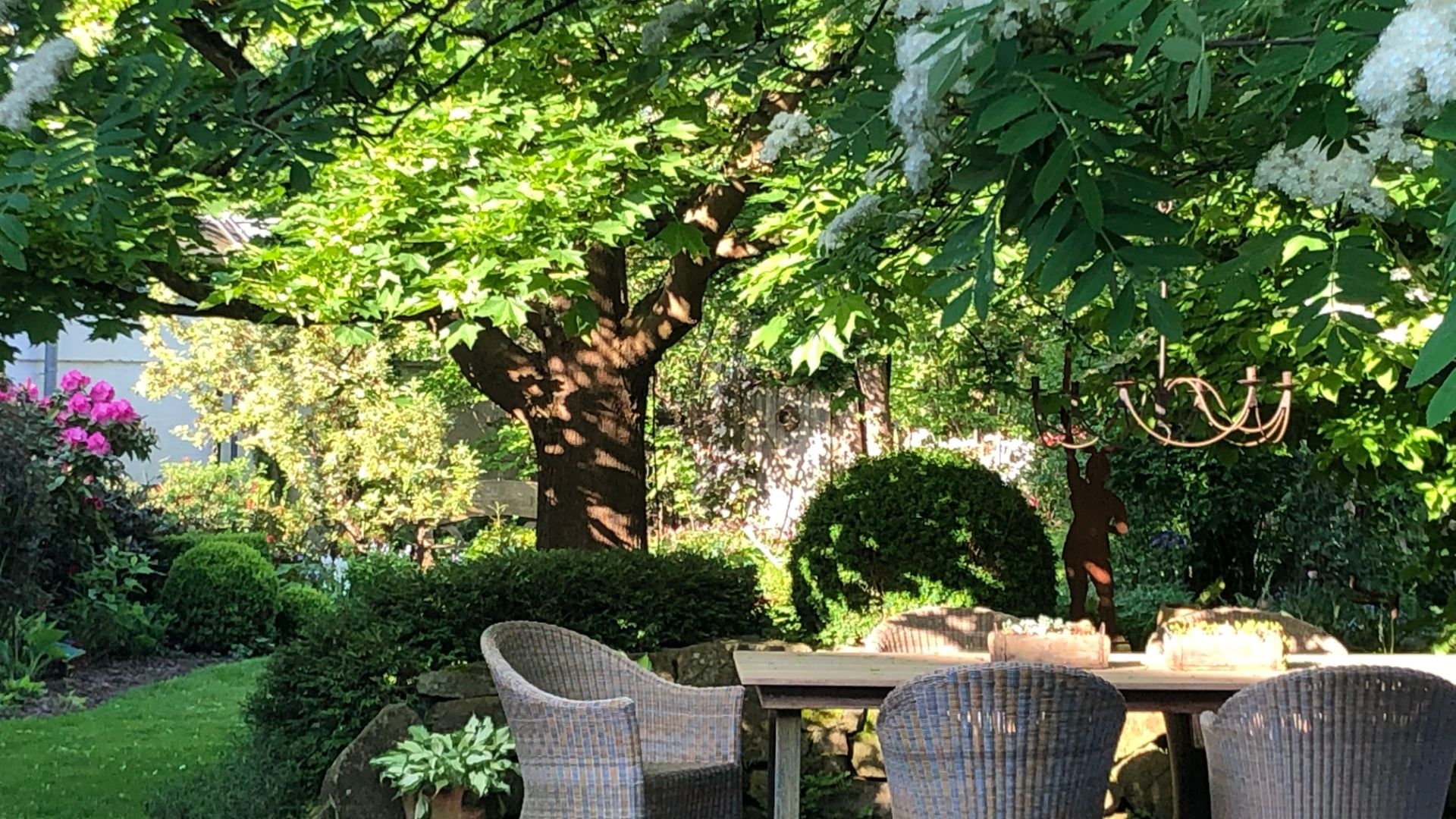 Sonniger Garten mit Holztisch, Korbstühlen, großen Bäumen, grünen Büschen, pinken Blumen und einer Silhouette
