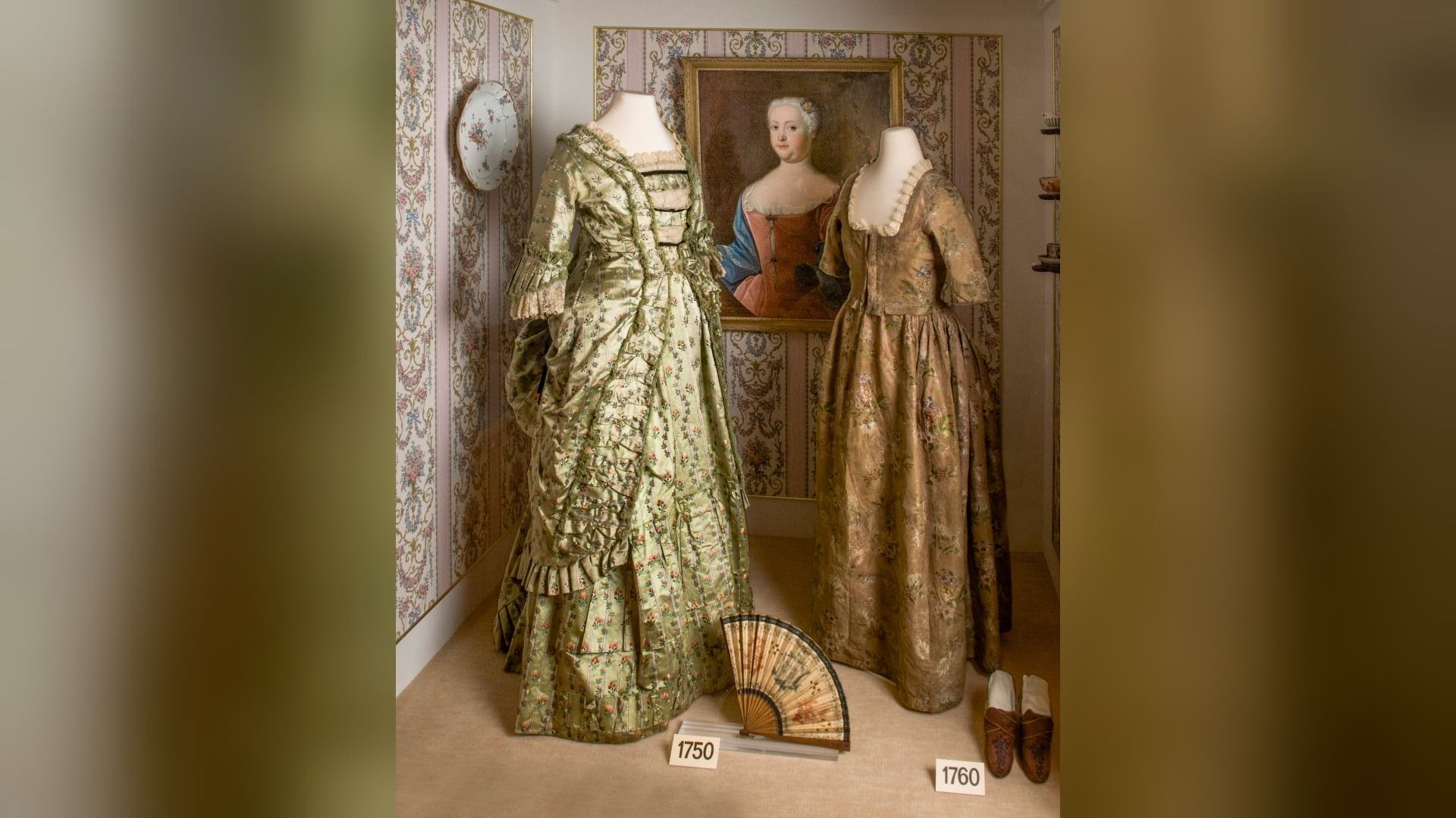 Zwei historische Kleider auf Puppen, ein Frauenportrait im Hintergrund, blumige Tapete, ein Fächer und Schuhe