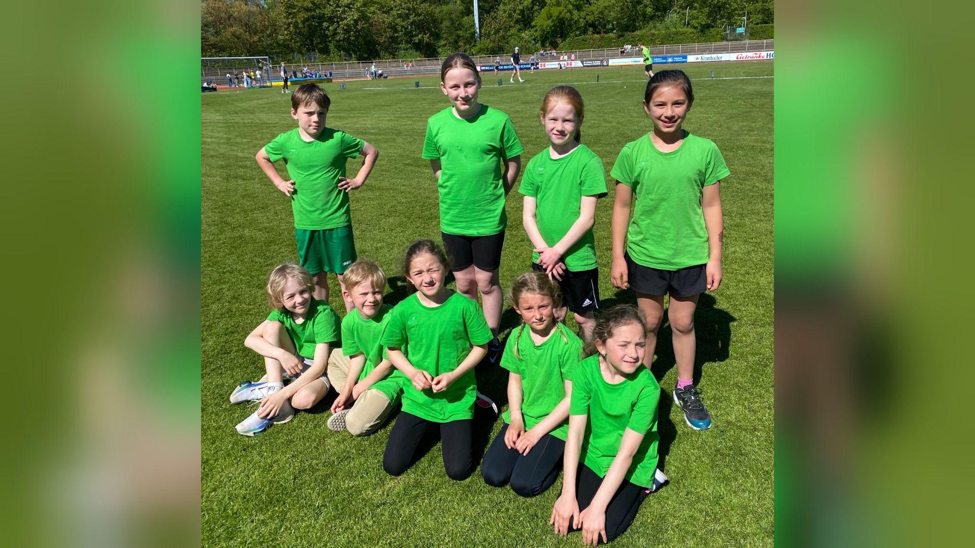 Neun Kinder in grünen T-Shirts stehen und knien lächelnd auf einem grünen Sportplatz unter blauem Himmel