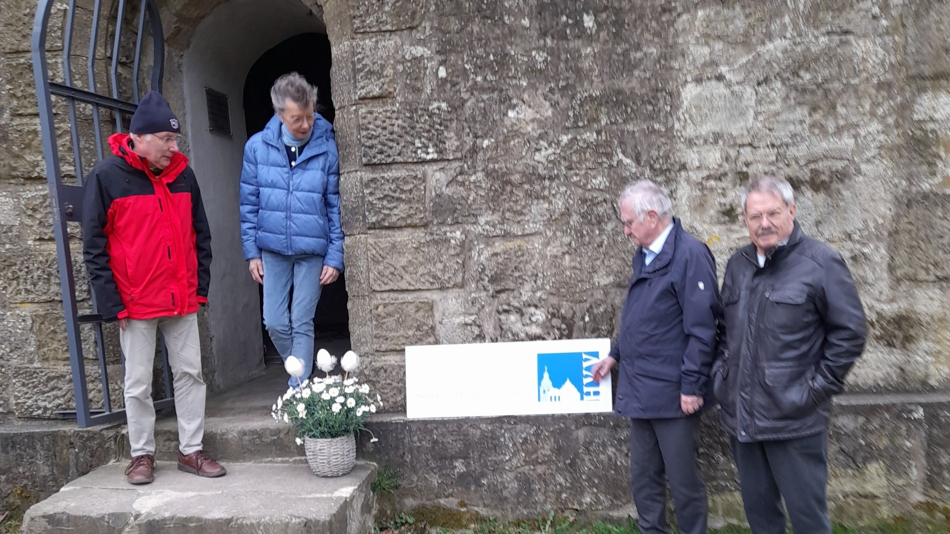 Vier Männer stehen am steinernen Eingang des Bismarckturms, davor ein weißes Schild mit blauem Logo und Blumen