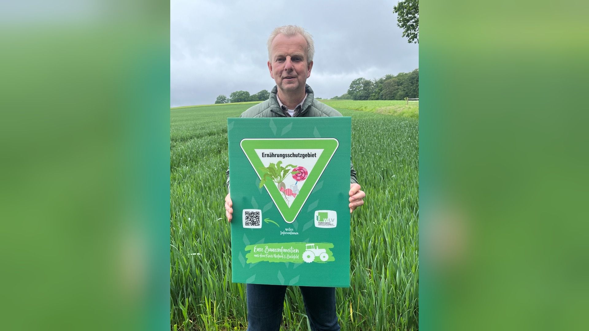Ein Mann steht in einem grünen Feld und hält ein Schild mit der Aufschrift „Ernährungsschutzgebiet“ und Abbildungen von