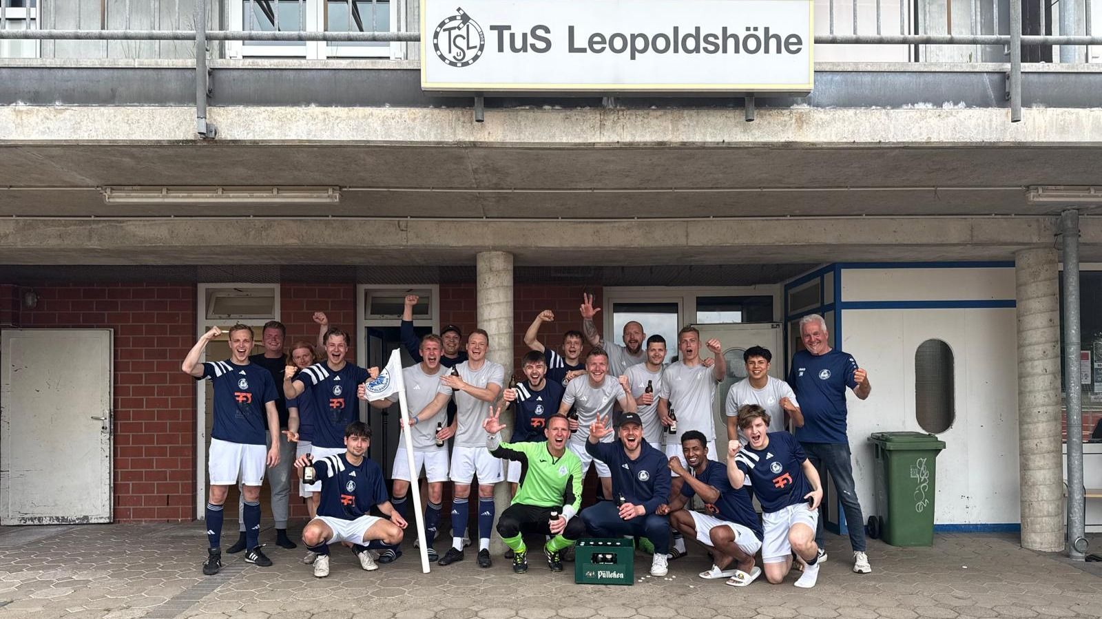 Eine jubelnde Fußballmannschaft in blauen und grauen Trikots feiert ihren Erfolg vor einem Vereinsgebäude mit