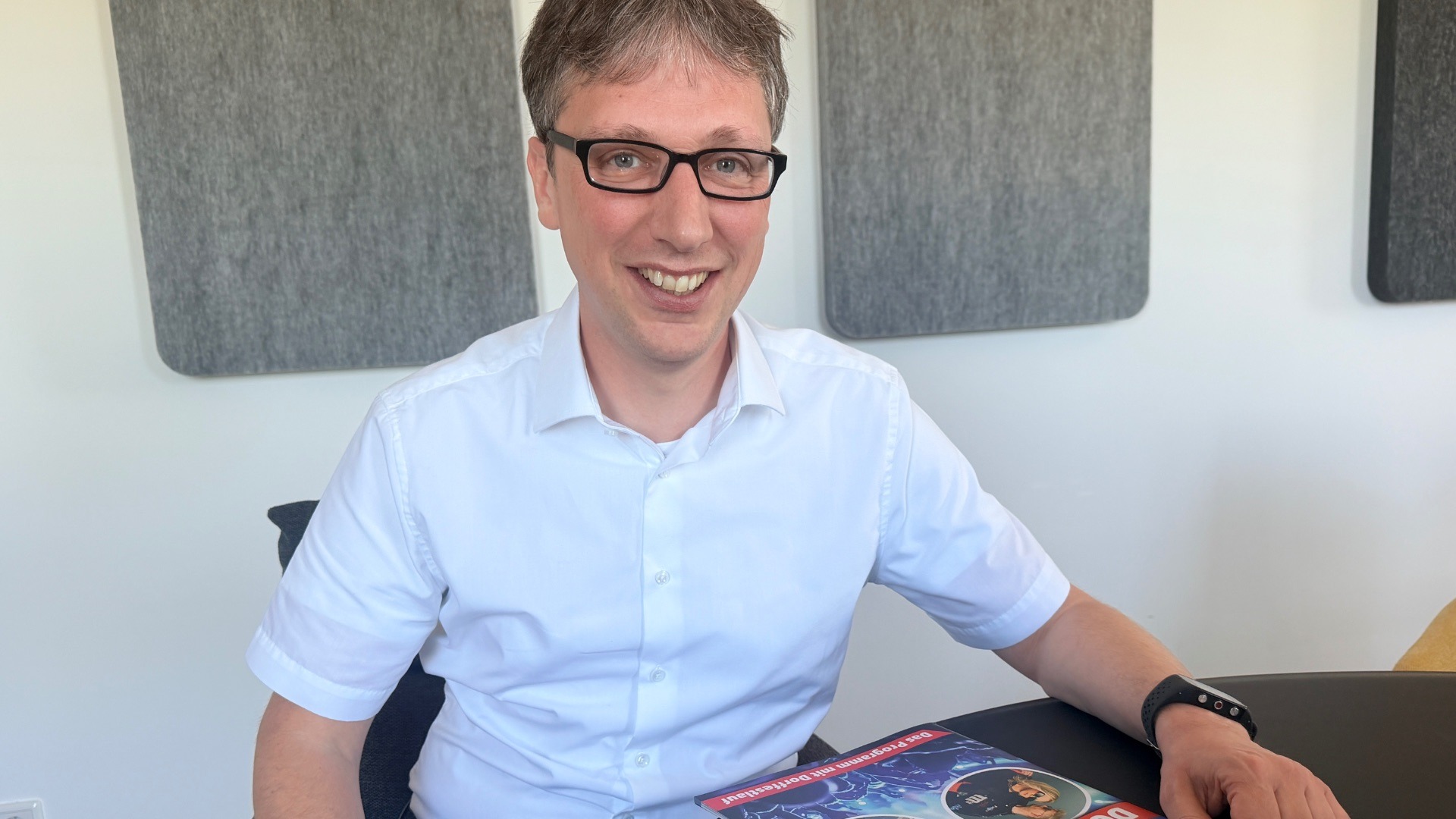 Bürgermeister Martin Hoffmann lächelt in die Kamera, sitzt am Tisch mit Kaffee und „Dorffest“-Magazin