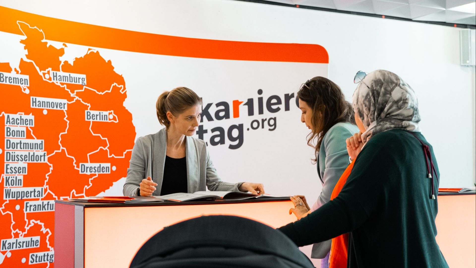 Eine Frau am Schreibtisch berät zwei Frauen, dahinter eine Deutschlandkarte mit Städten und "karrieretag.org