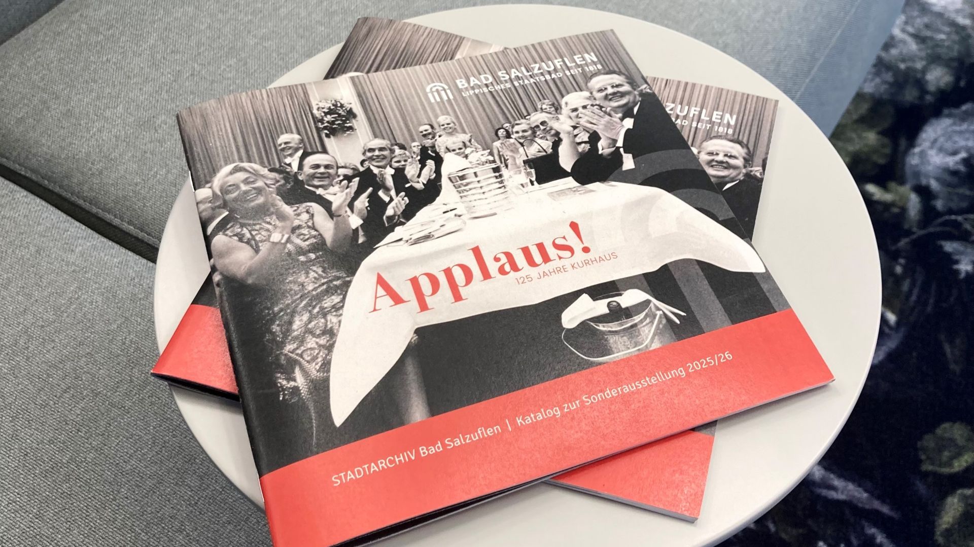 Mehrere Kataloge „Applaus! 125 Jahre Kurhaus Bad Salzuflen“ mit klatschenden Menschen auf dem Cover auf einem Tisch