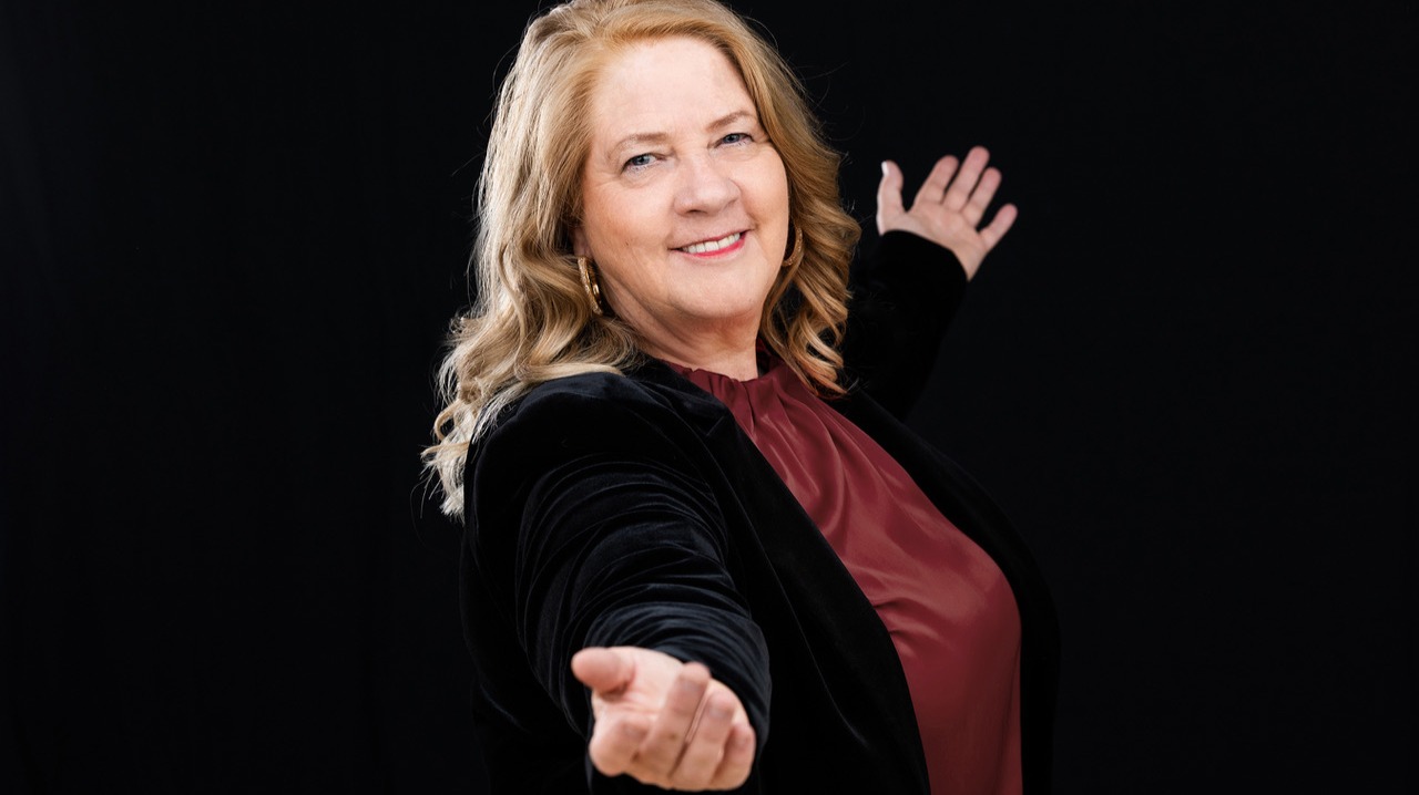 Kathy Kelly lächelt und gestikuliert mit beiden Armen einladend vor schwarzem Hintergrund