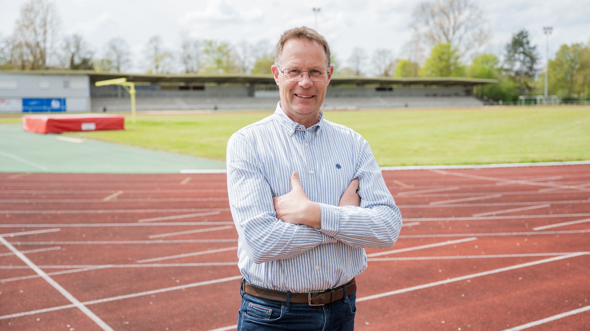 Lächelnder Mann mit Brille und verschränkten Armen auf roter Leichtathletikbahn vor Tribüne und Bäumen