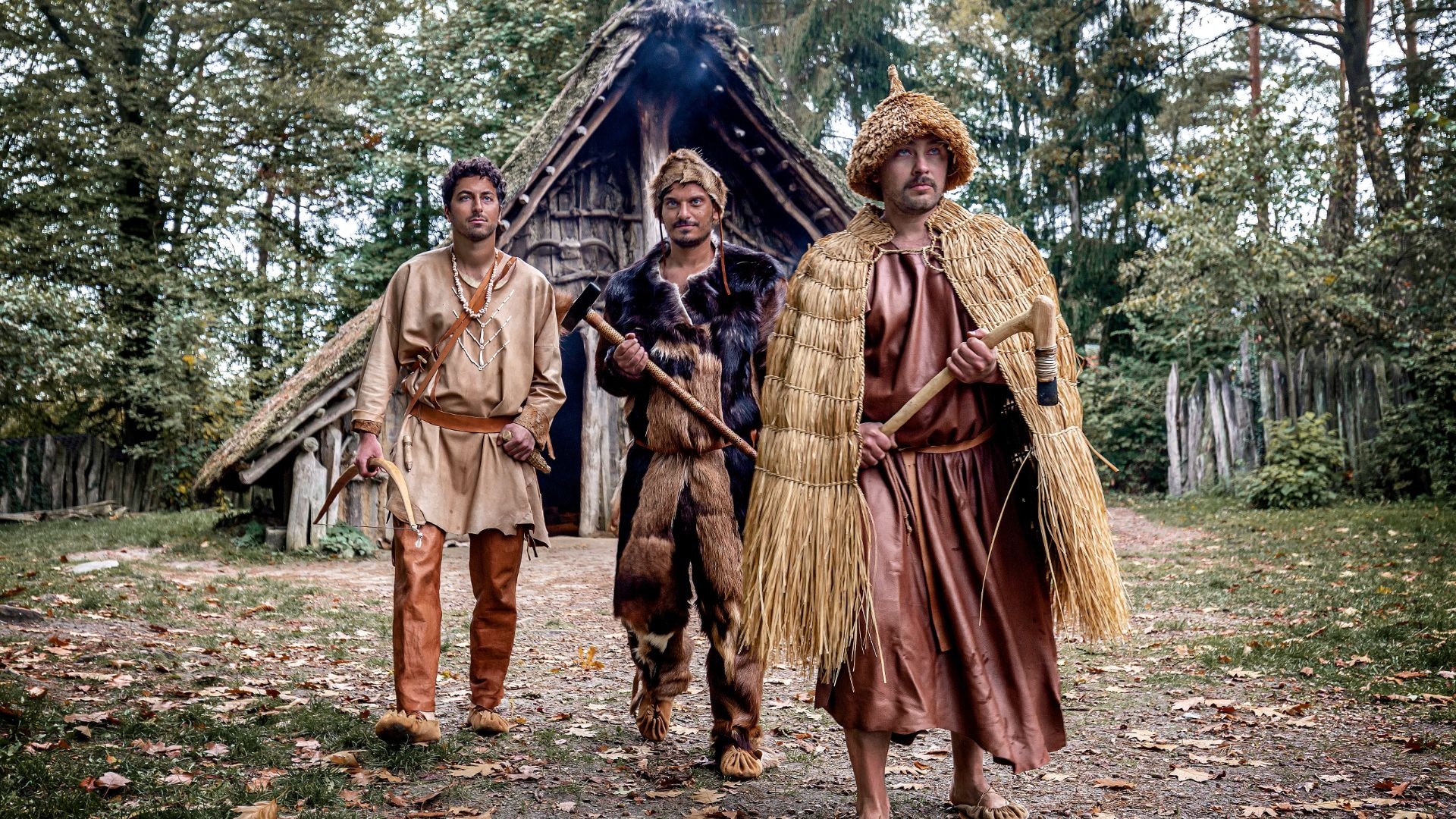 Drei Männer in Steinzeitkleidung mit Bogen und Äxten vor einer Holzhütte im Freilichtmuseum Oerlinghausen