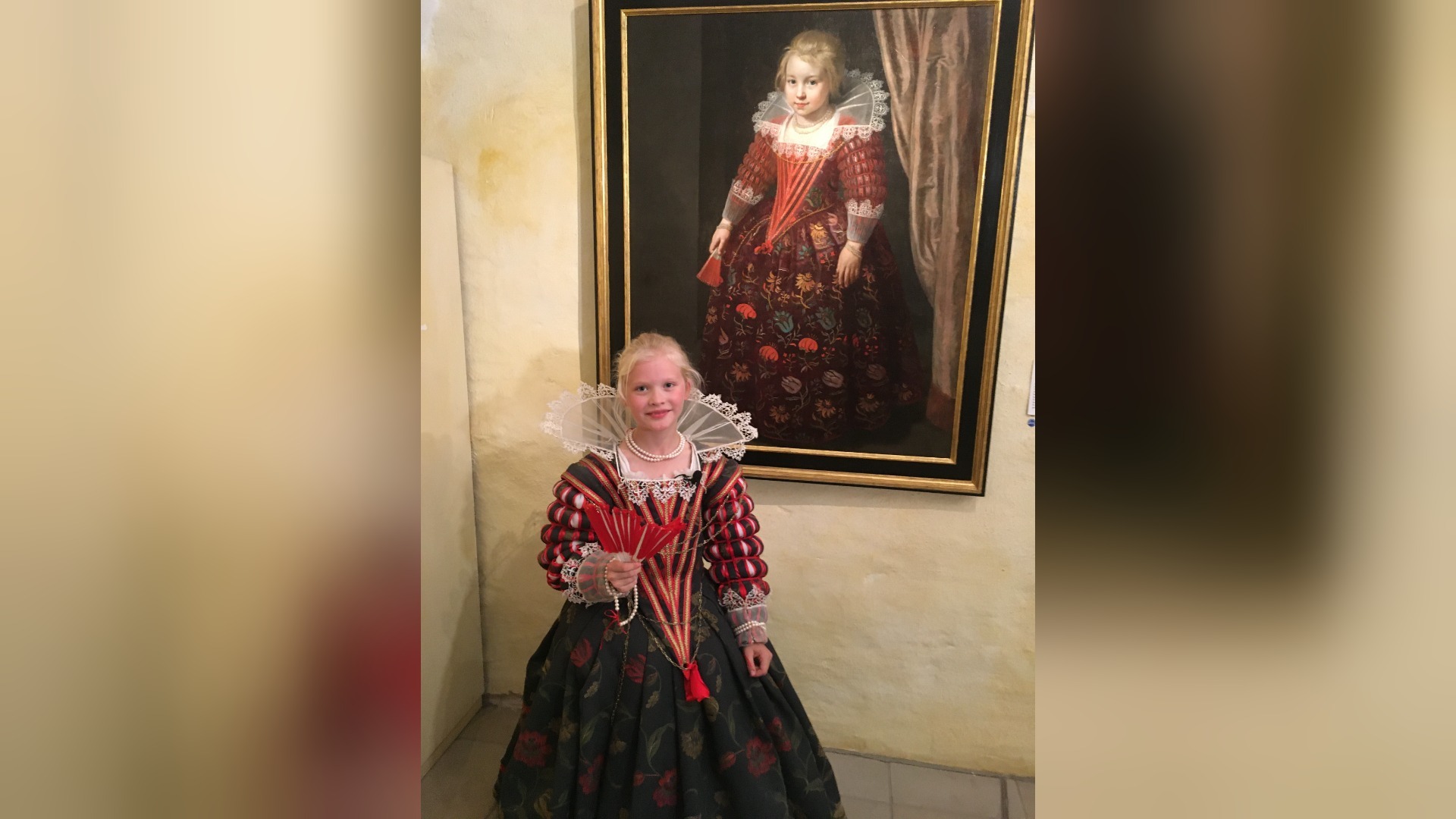 Blondes Mädchen in roter Renaissancetracht mit Fächer steht vor einem Gemälde eines Mädchens in ähnlichem Kleid