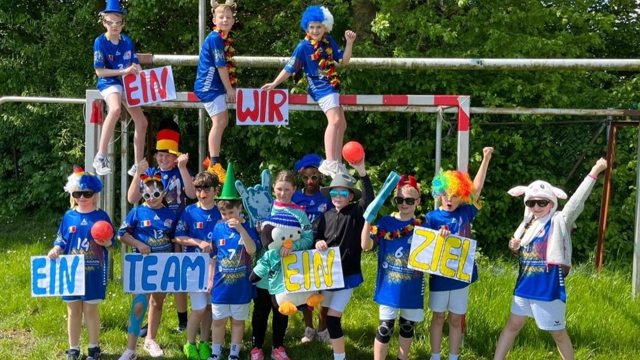 Viele Kinder eines Handballteams in blauen Trikots, festlichen Verkleidungen und Schildern "Ein Team Ein Ziel