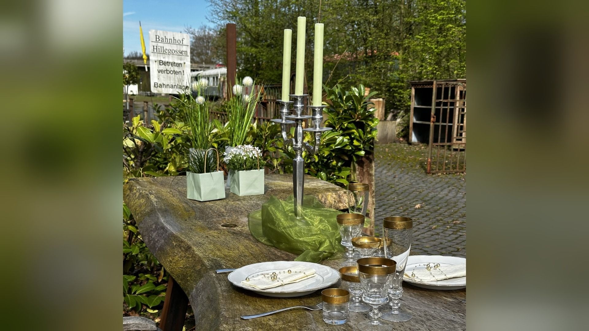 Gedeckter Holztisch im Freien mit silbernem Kerzenständer, grünen Kerzen, Tellern, Gläsern und Pflanzen vor einem