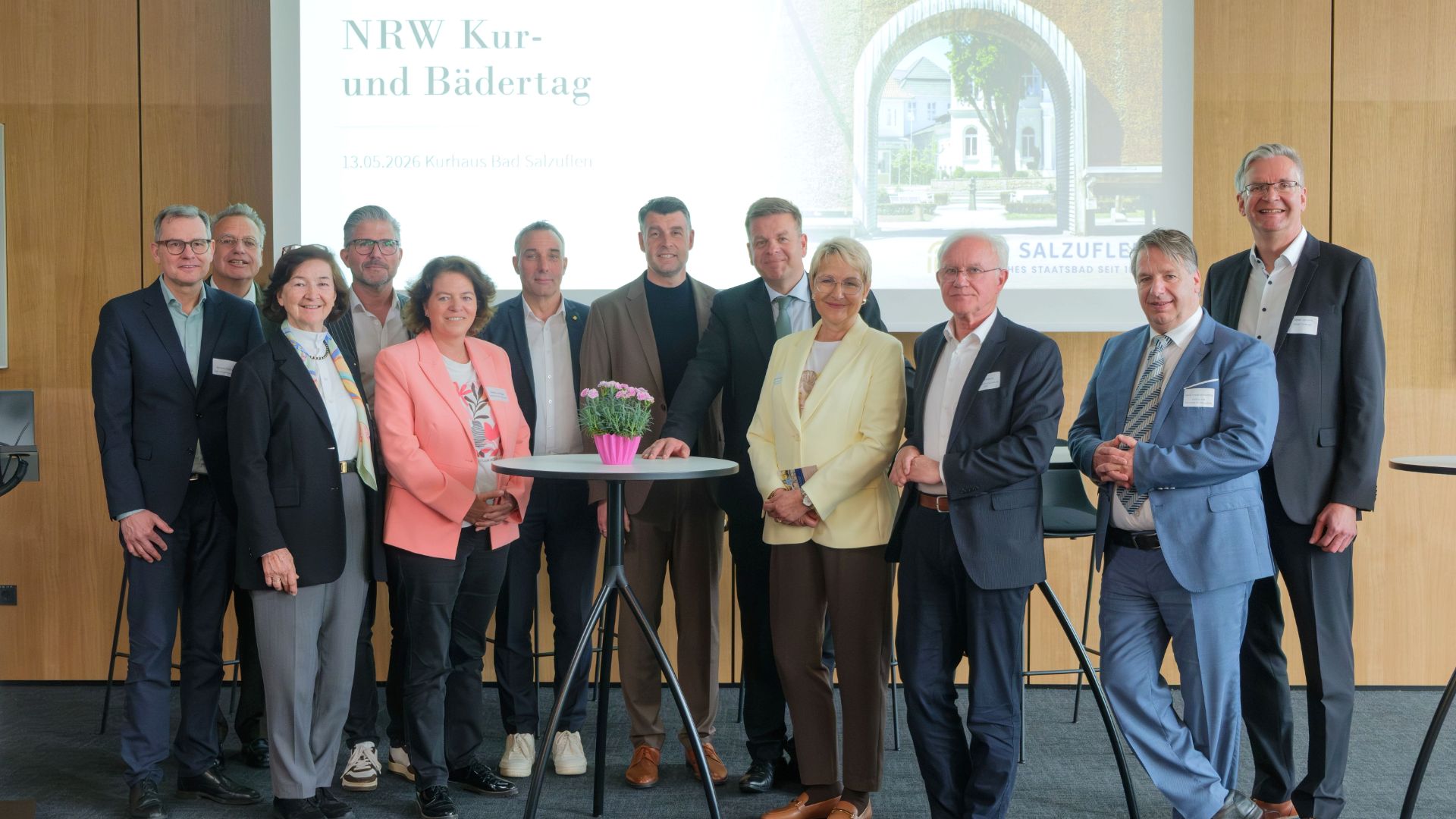 13 Männer und Frauen stehen in einer Gruppe vor einer Projektionsfläche mit 'NRW Kur- und Bädertag' und einem Stehtisch