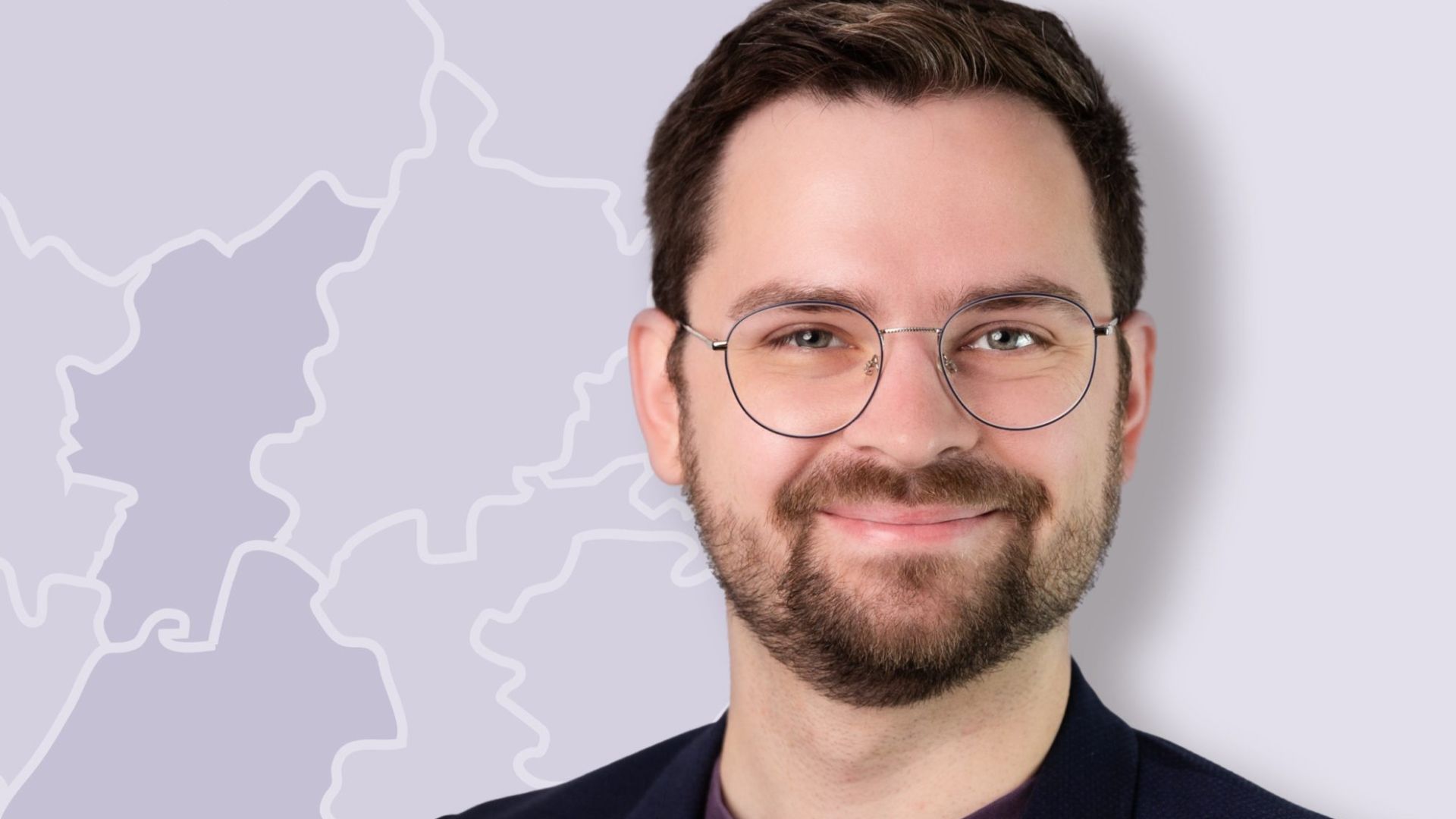 Lächelnder Mann mit Bart und Brille im Sakko vor einer schematischen Landkarte