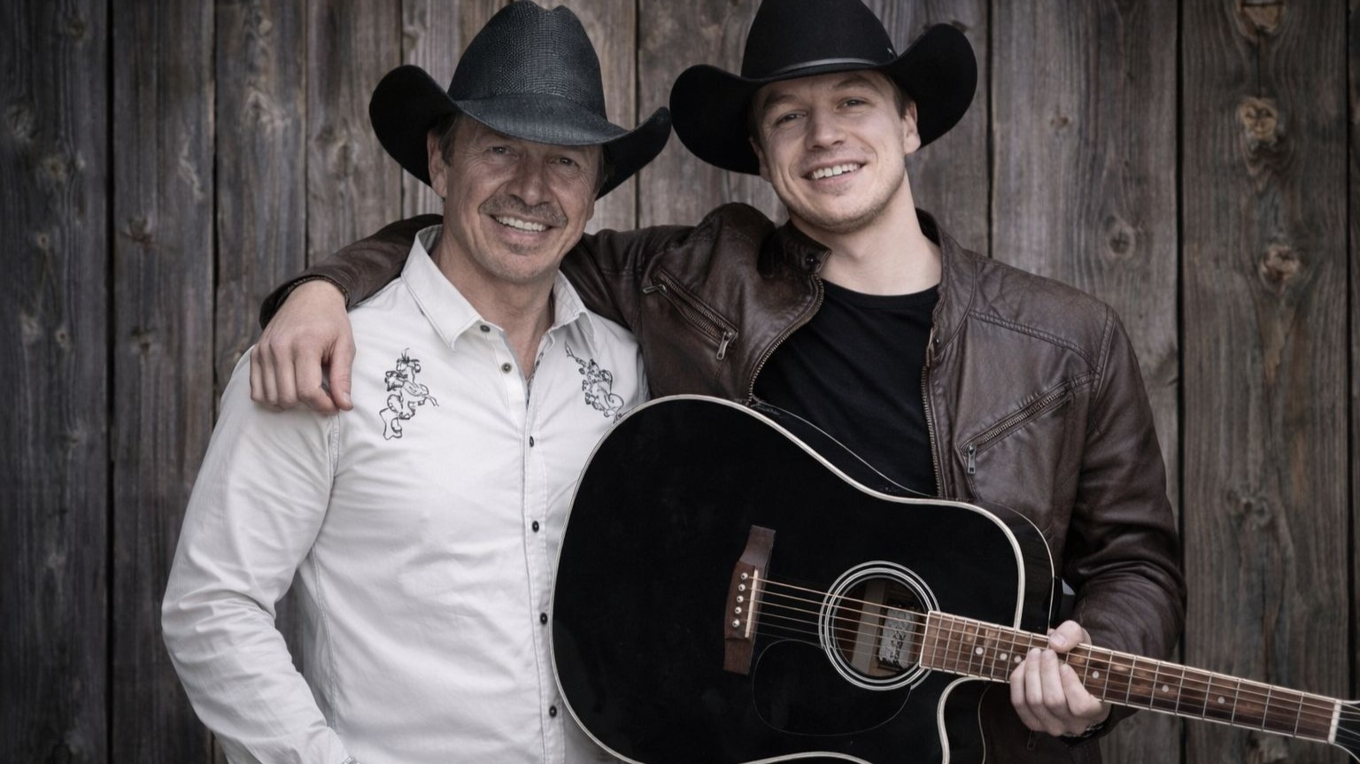 Zwei lächelnde Männer in Cowboyhüten, der eine mit Arm um den anderen, hält eine schwarze Akustikgitarre vor einer