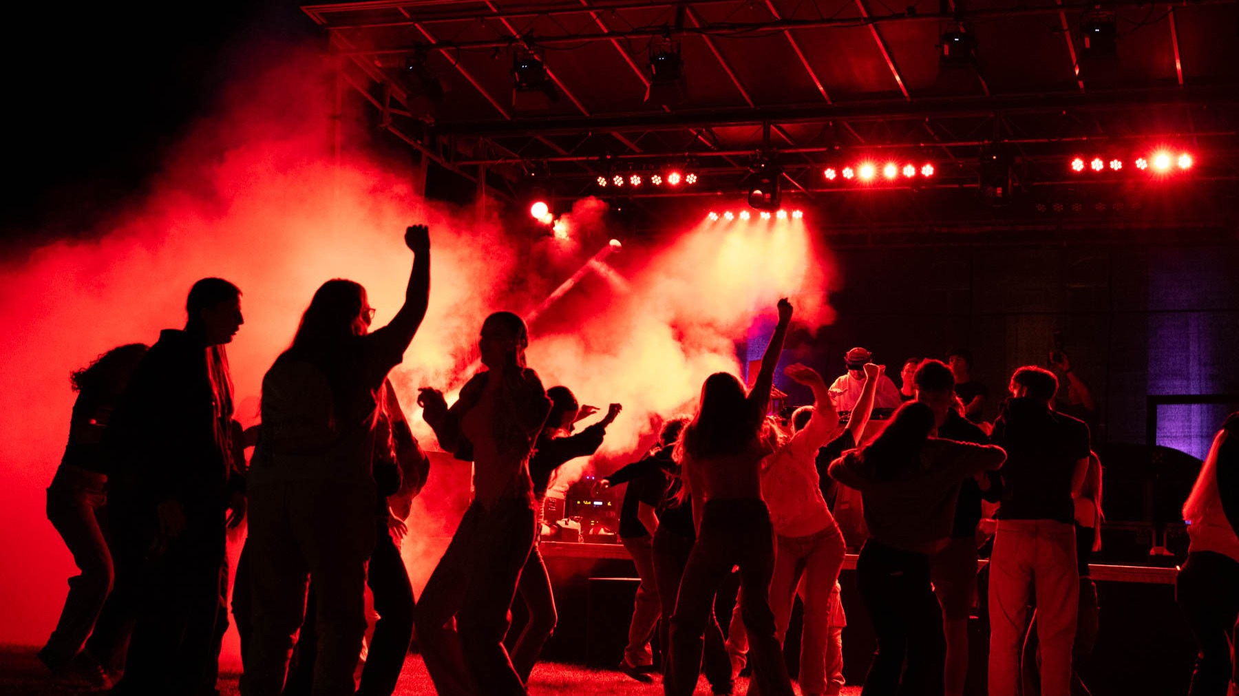 Zahlreiche Feiernde tanzen im roten Licht und Nebel vor einer beleuchteten Bühne auf einer Party
