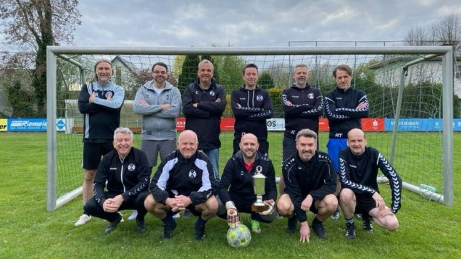 Zehn Männer in Sportkleidung posieren mit einem Pokal und Fußball vor einem Fußballtor auf einem Sportplatz