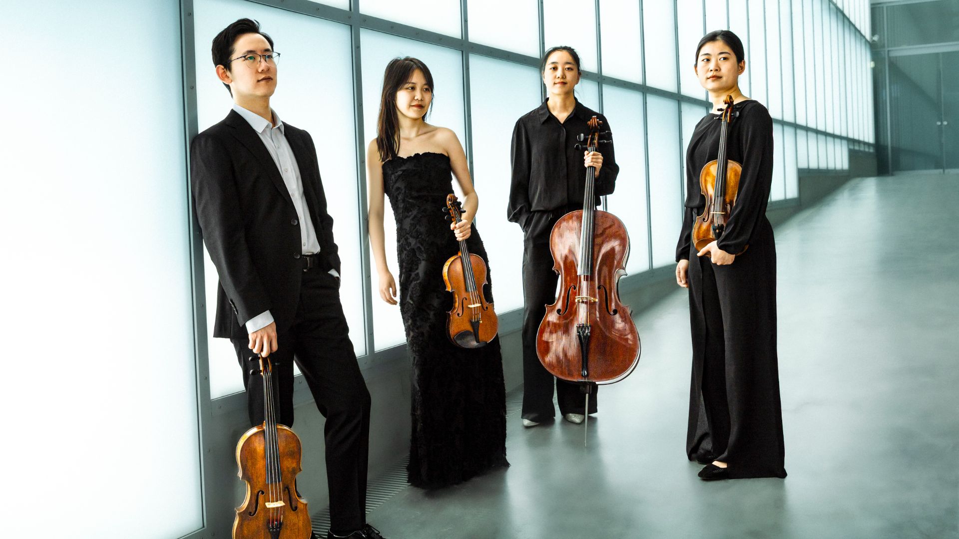 Ein Mann und drei Frauen als Musiker stehen in Abendkleidung mit drei Violinen und einem Cello vor einer Glasfassade