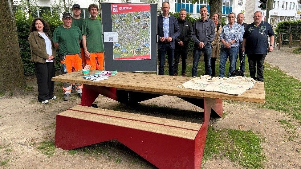Elf Personen stehen hinter einem roten Holztisch und einer Stadtplan-Tafel in einem Park in Hillegossen