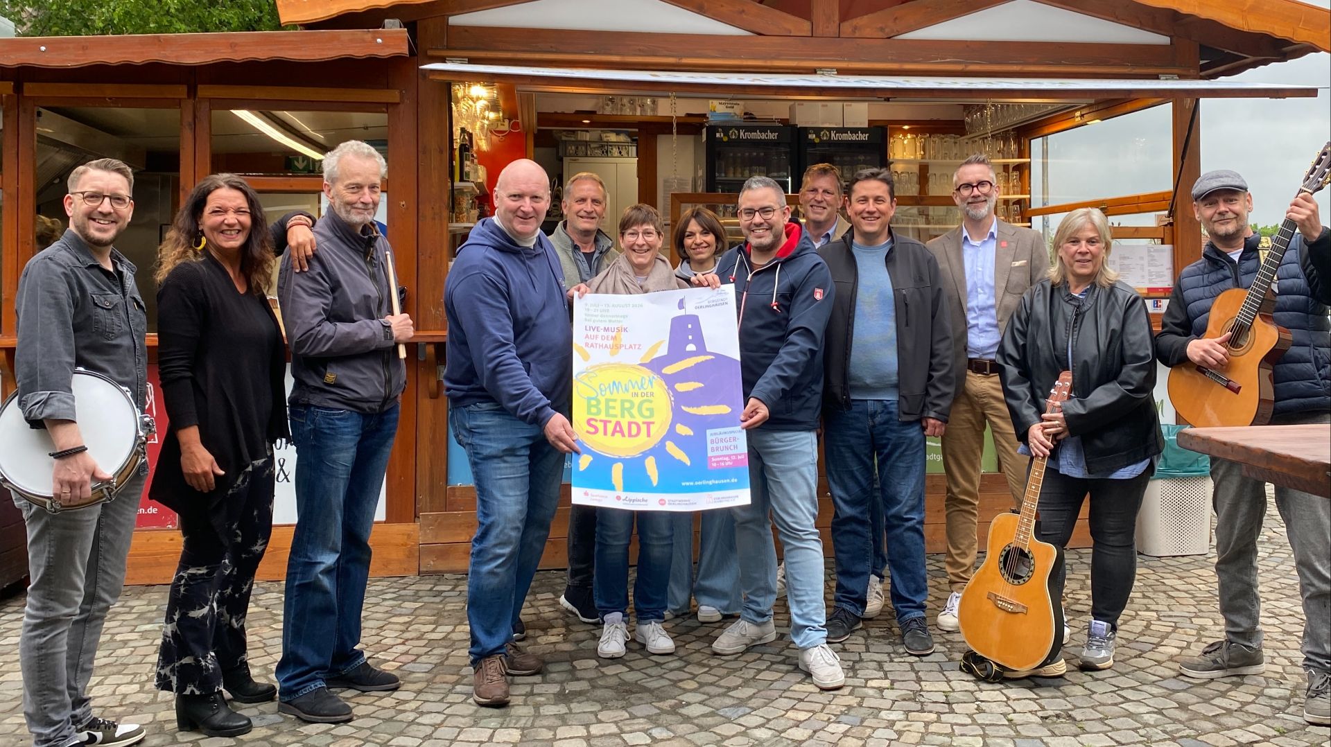 Gruppe von 15 Personen mit Instrumenten präsentiert Plakat für Oerlinghausener Sommerfest vor Verkaufsstand