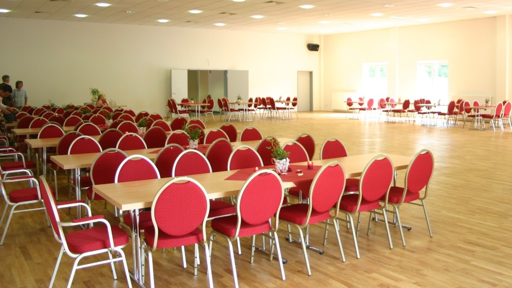 Heller Tanzsaal mit Parkettboden, vielen Tischen und roten Stühlen in Reihen aufgestellt