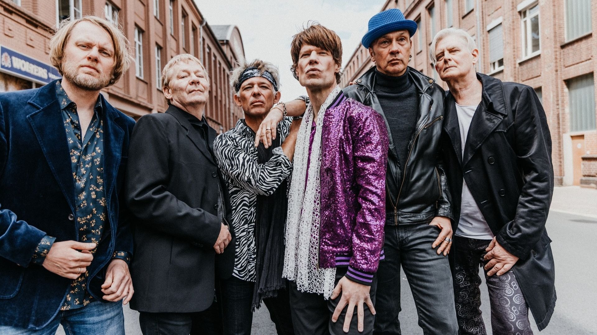 Sechs Musiker der Band Voodoo Lounge in Bühnenkostümen posieren auf einer Straße vor Backsteingebäuden