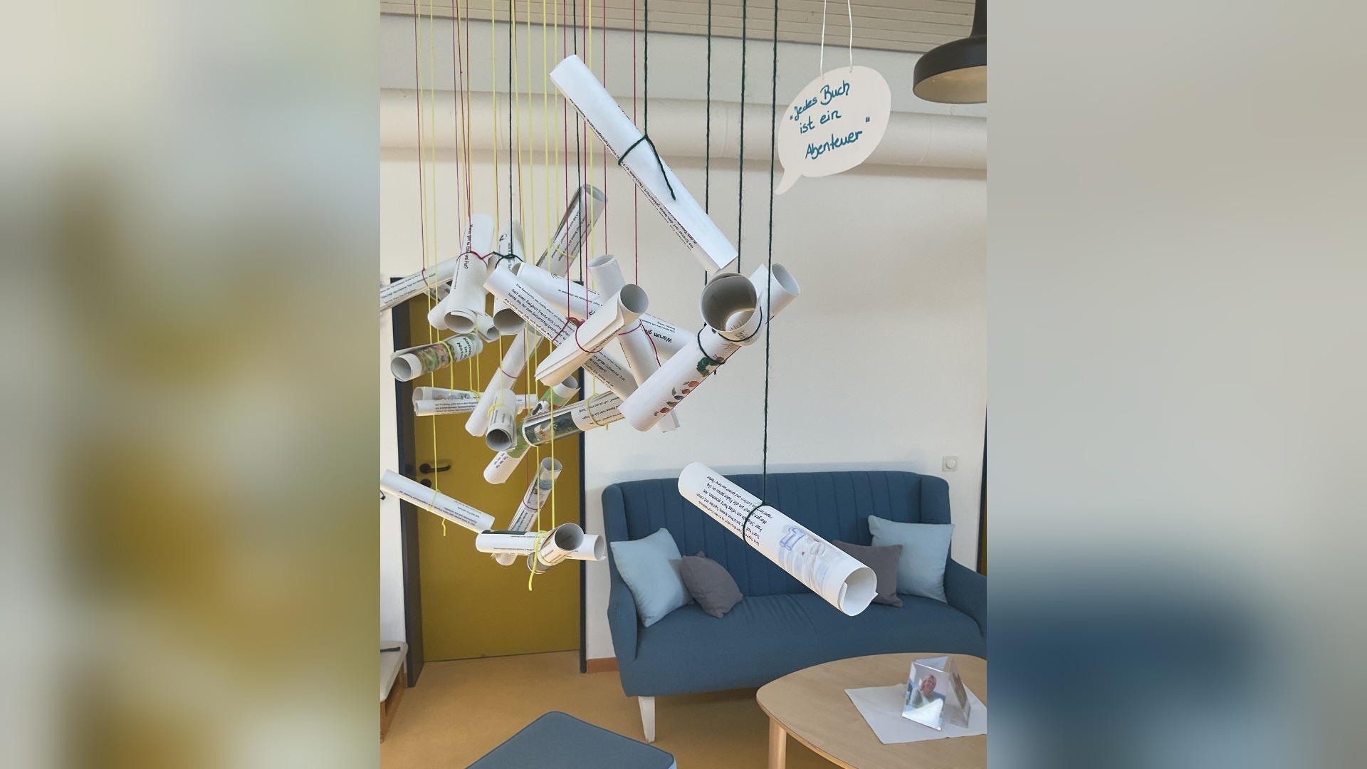 Gerollte Papiere hängen als Geschichten-Regen von der Decke über einem Sofa, ein Schild sagt "Jedes Buch ist ein