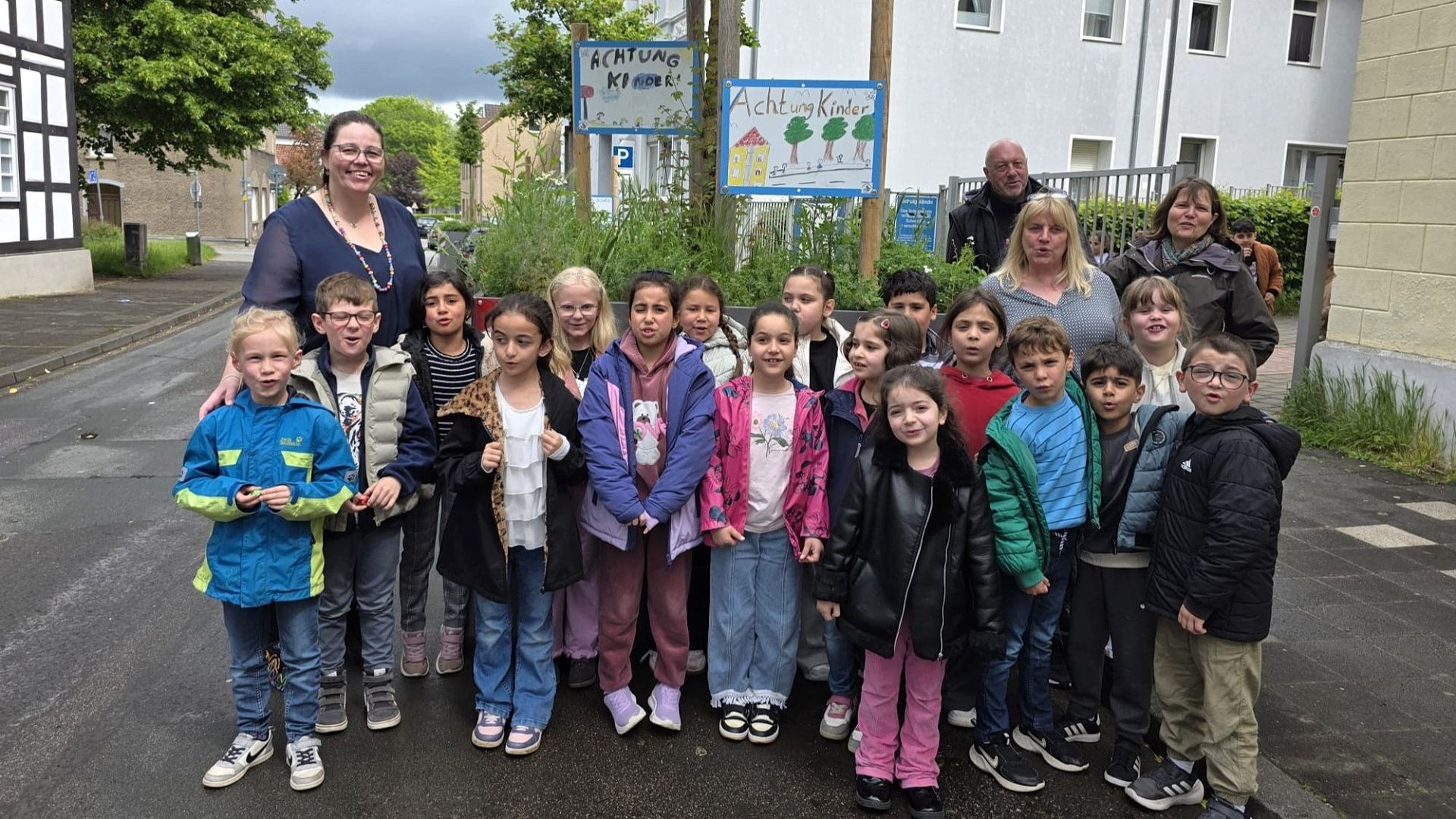 Eine Gruppe von etwa 20 Grundschulkindern und 4 Erwachsenen steht auf einer Straße vor handgemachten „Achtung