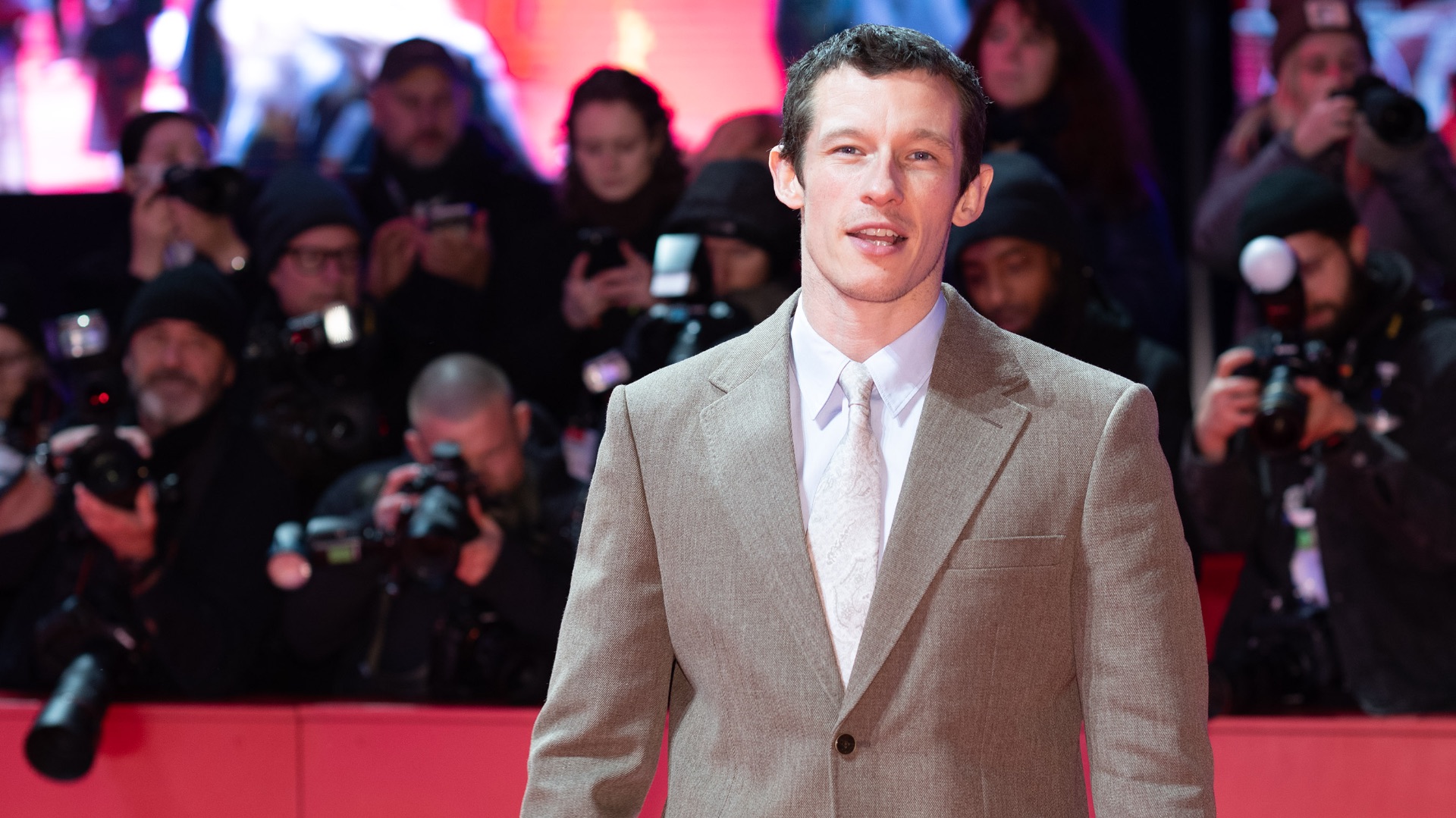 Callum Turner in hellem Anzug auf dem roten Teppich, umringt von Fotografen