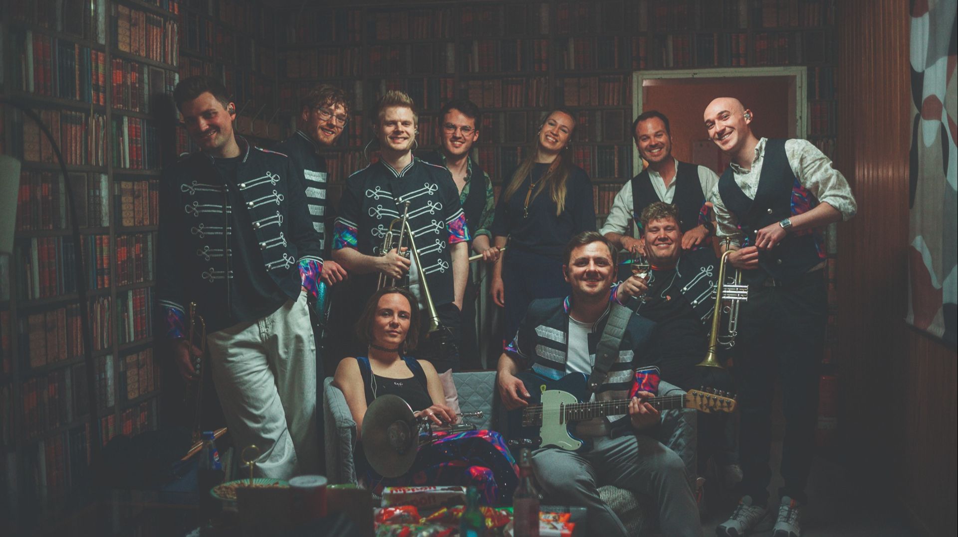 Zehn lächelnde Musiker der Band Druckluft posieren mit Instrumenten in einem Raum vor einem Bücherregal