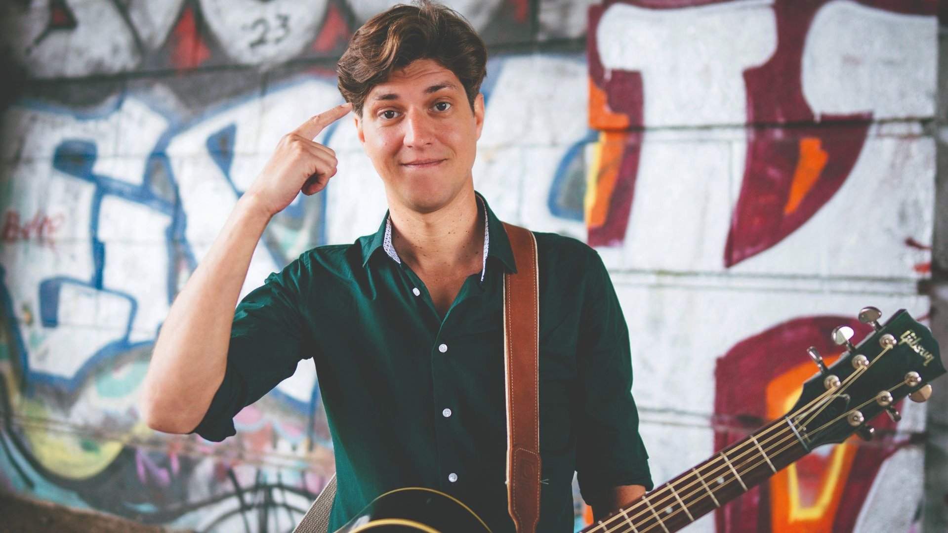 FALK mit Gitarre zeigt mit dem Finger an die Schläfe und lächelt vor einer bunten Graffitiwand