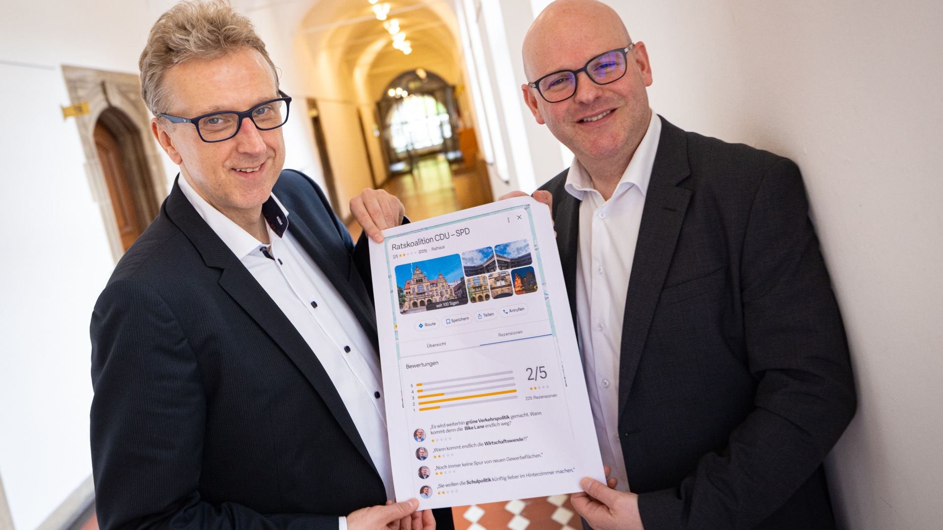 Zwei lächelnde Männer halten ein Blatt Papier mit der Bewertung „Ratskoalition CDU-SPD 2/5 Sterne“ hoch