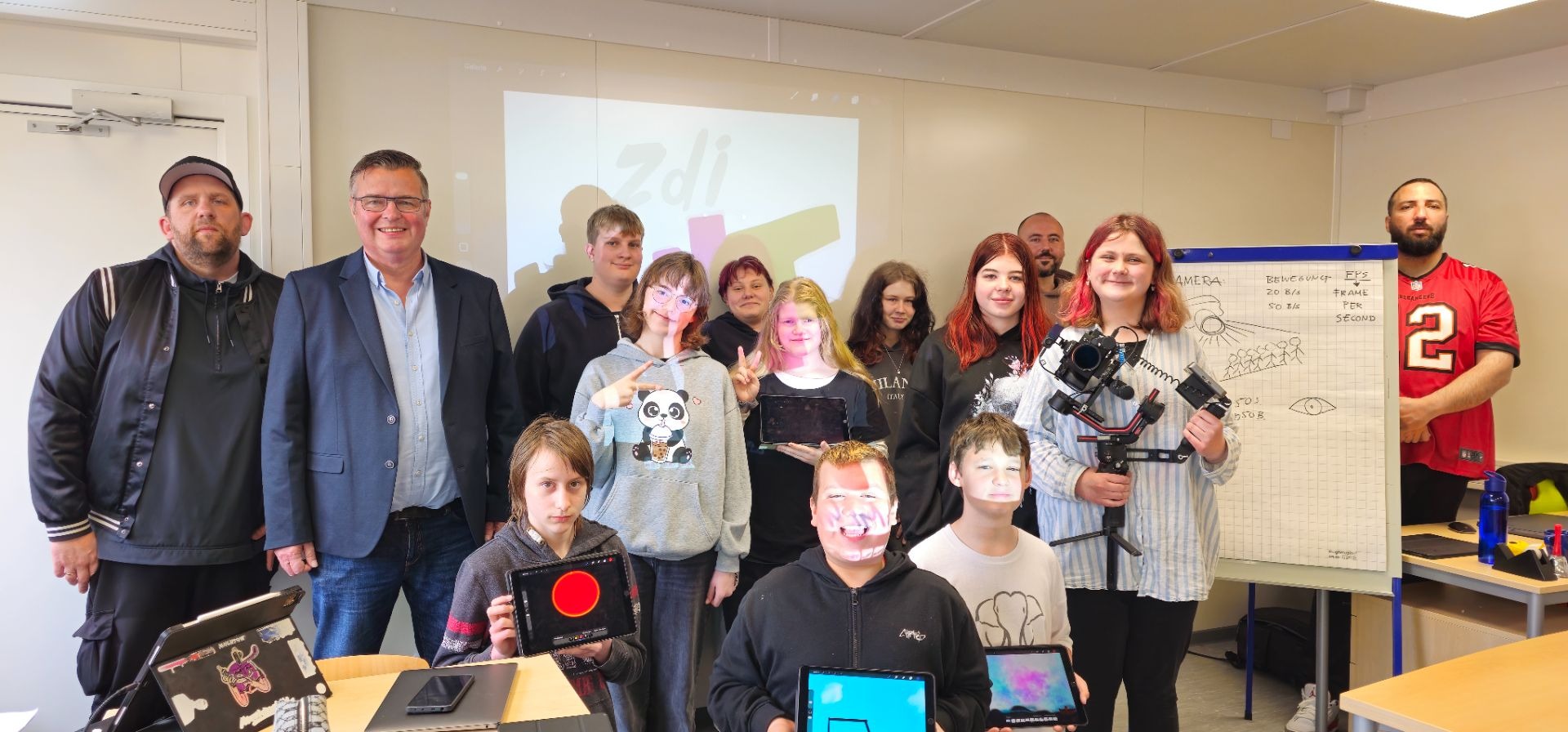Gruppe von Schülern und drei Erwachsenen lächelt in einem Lernraum, manche halten Tablets und eine Videokamera,