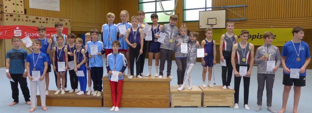 Junge Turnerinnen und Turner mit Medaillen und Urkunden stehen auf einem Siegerpodest in einer Sporthalle