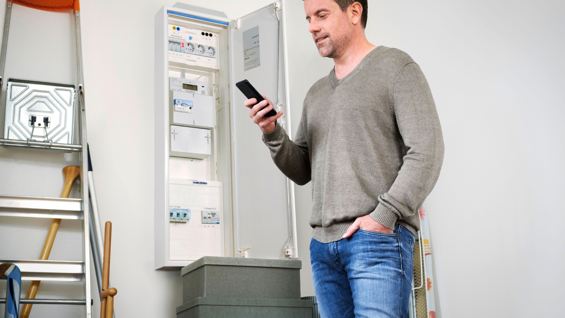Mann schaut auf sein Smartphone neben geöffnetem Stromkasten mit Zähler