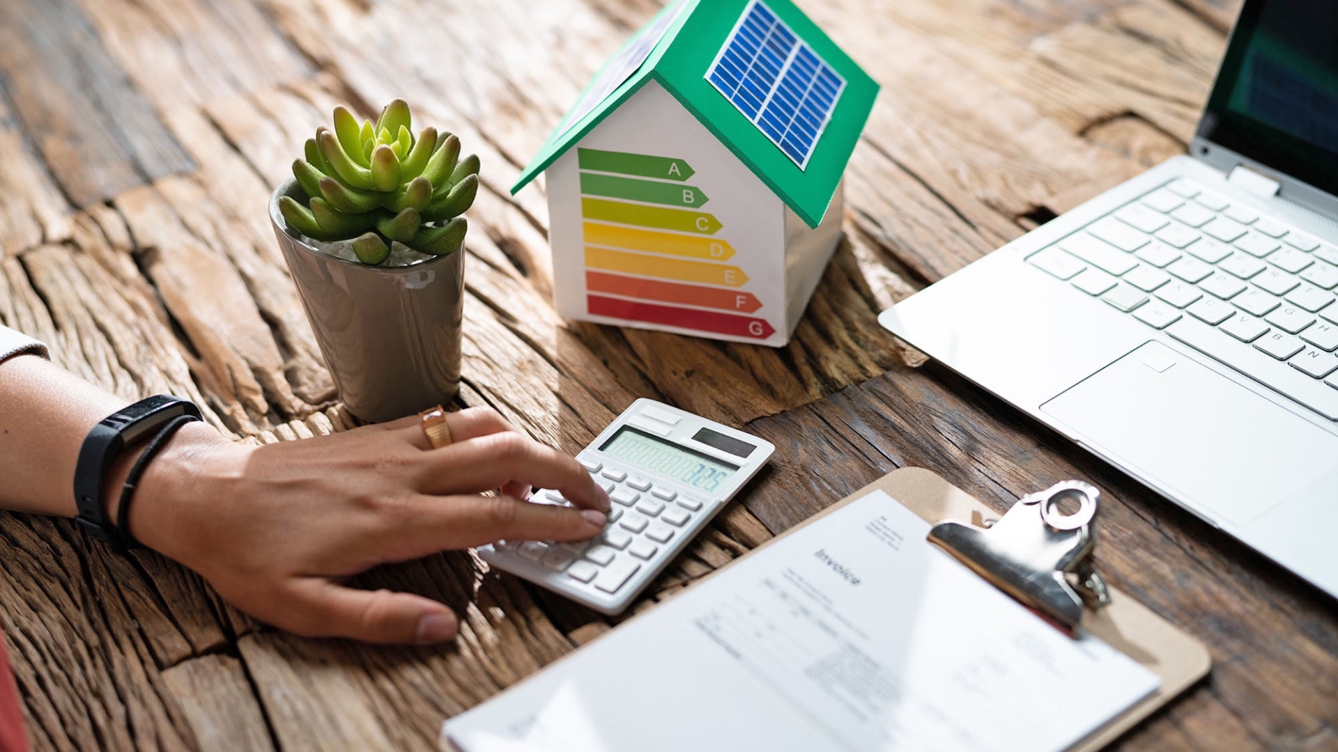 Hand rechnet auf Taschenrechner neben Hausmodell mit Solardach, Energieeffizienzskala, Laptop und Rechnung auf Holztisch