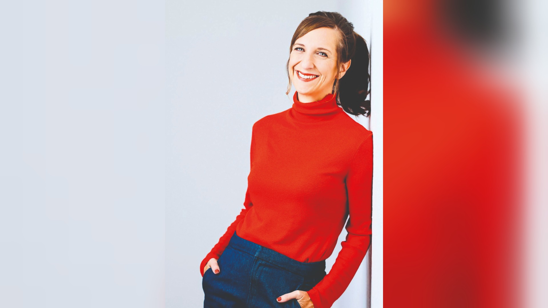 Sarah Hakenberg lächelt in rotem Rollkragenpullover und blauer Hose, Hände in den Taschen, an helle Wand gelehnt