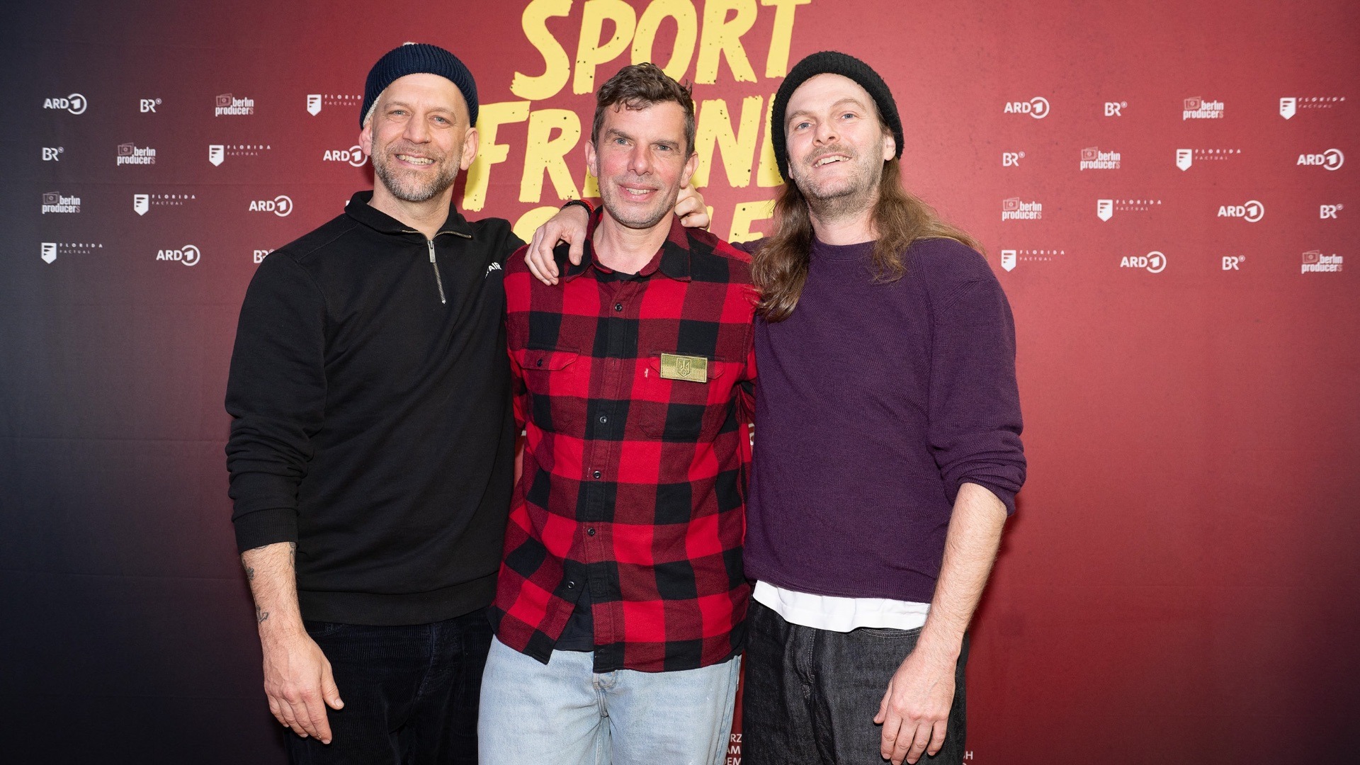 Drei Männer der Band Sportfreunde Stiller lächeln vor einer roten Wand mit Logo-Prints und Schriftzug
