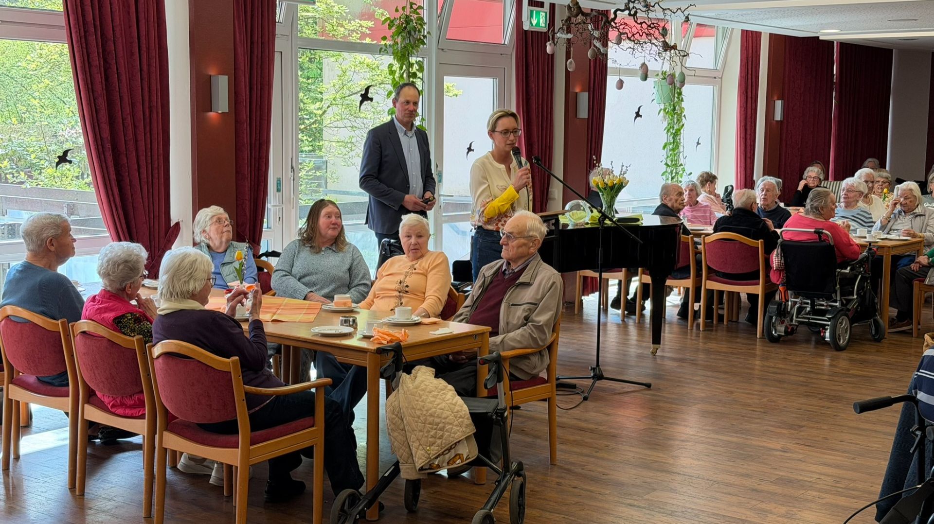 Eine Frau spricht am Mikrofon vor einer Gruppe Senioren, die in einem hellen Raum an Tischen sitzen und zuhören