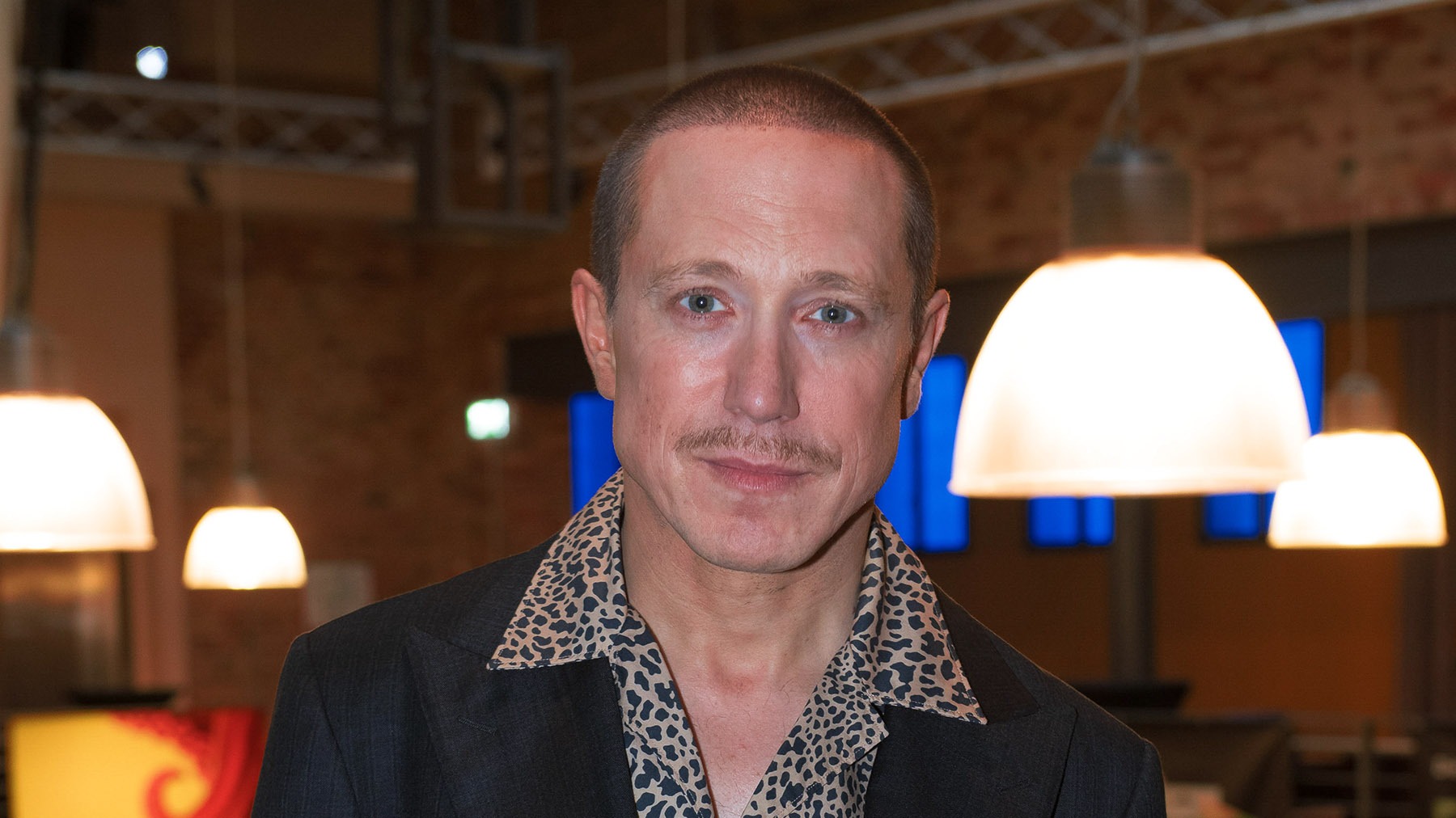 Timo Jacobs mit kurzen Haaren, Schnurrbart und gemustertem Hemd blickt frontal, im Hintergrund leuchtende Lampen