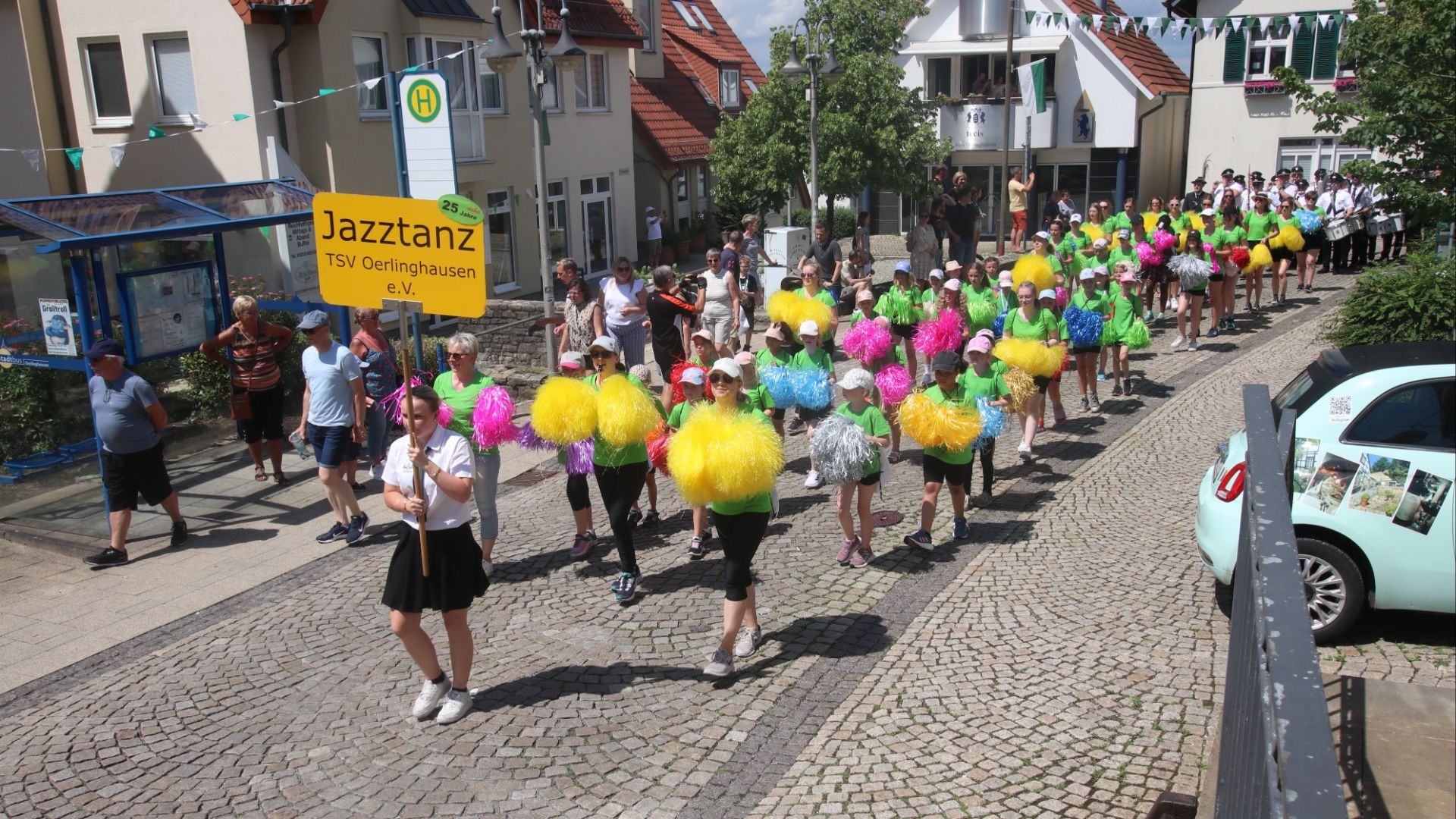 Ein Festumzug mit Jazztanzgruppe und Blaskapelle zieht auf einer gepflasterten Straße durch eine Stadt, Zuschauer am