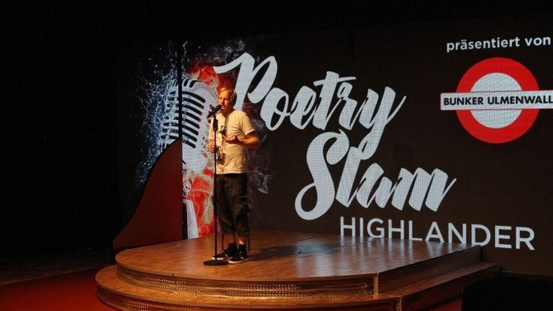 Junger Mann spricht an Mikrofon auf Holz-Bühne vor Leinwand mit "Poetry Slam Highlander" und "BUNKER ULMENWALL