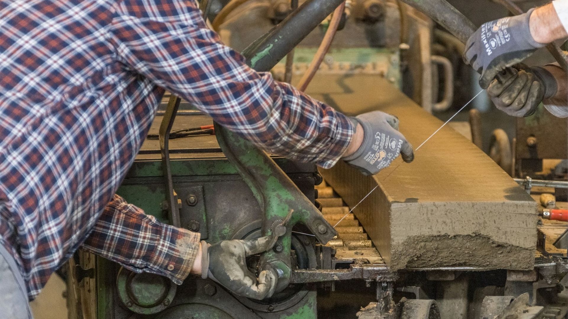 Hände in Handschuhen schneiden eine lange, feuchte Lehmmasse mit einem Draht auf einer Ziegelmaschine