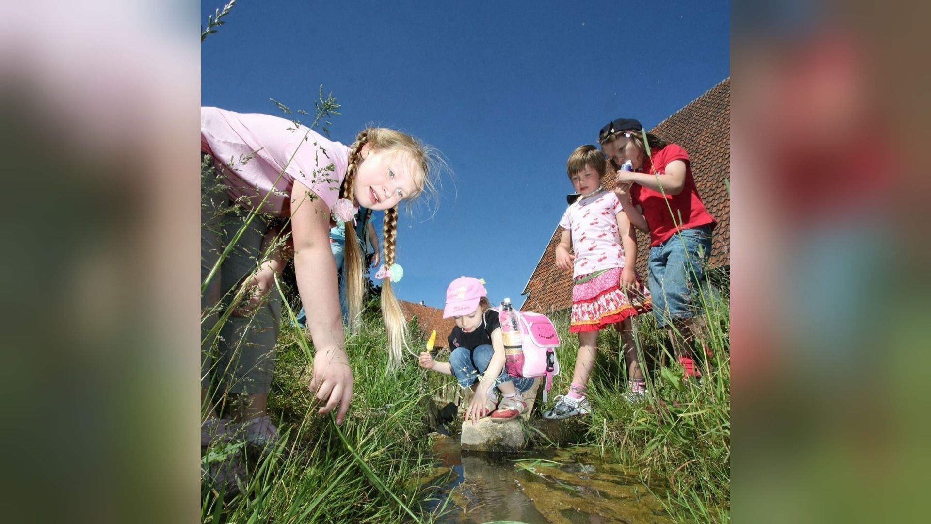 Mehrere Kinder spielen an einem kleinen Bach im Gras unter blauem Himmel, ein Mädchen blickt in die Kamera