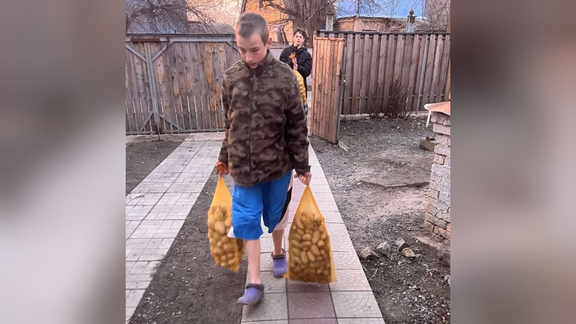 Junger Mann trägt zwei Netze voller Kartoffeln auf einem gepflasterten Weg, dahinter geht eine weitere Person