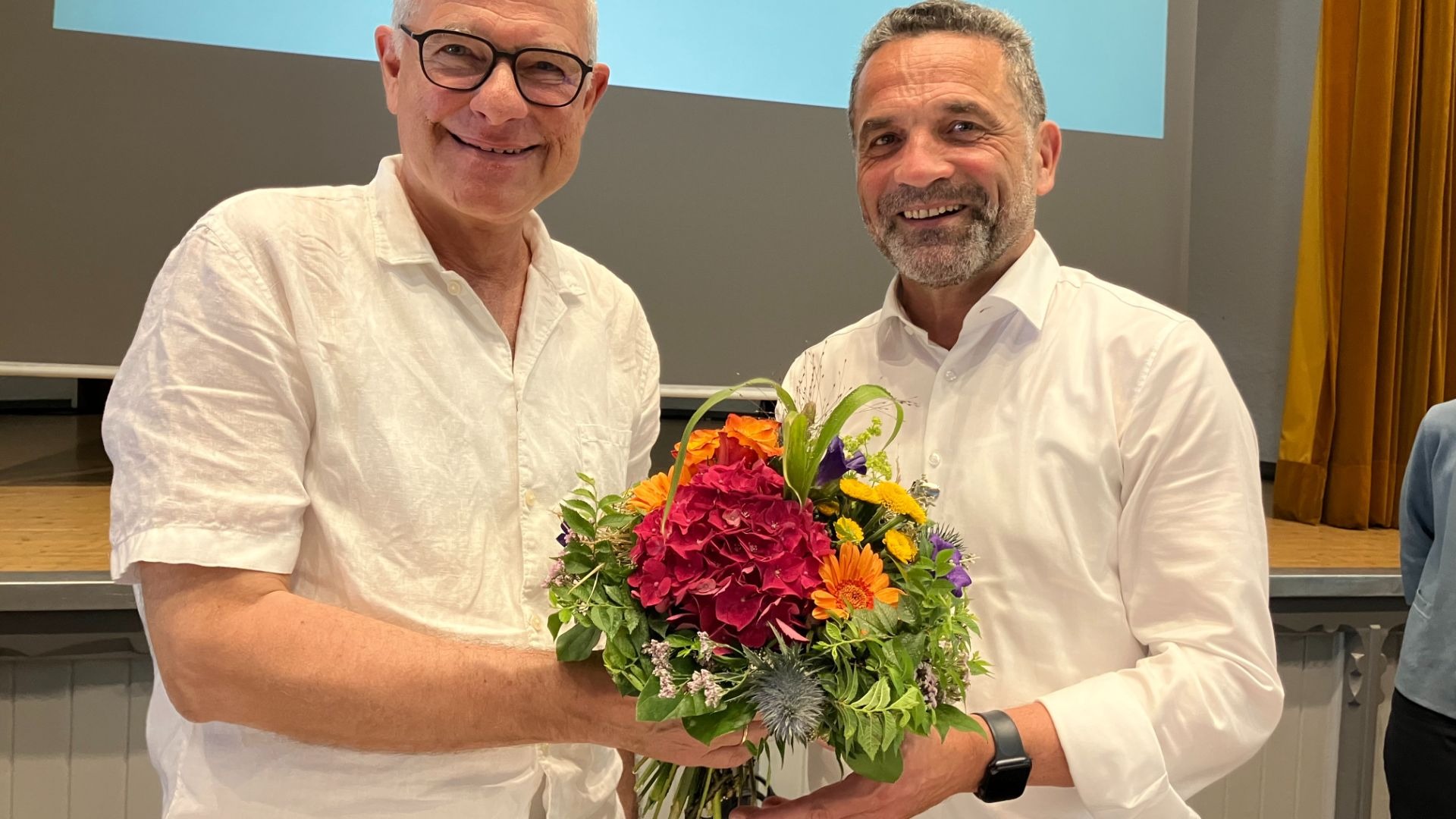 Zwei lächelnde Männer in weißen Hemden, einer mit Brille, der andere bärtig, halten einen farbenfrohen Blumenstrauß