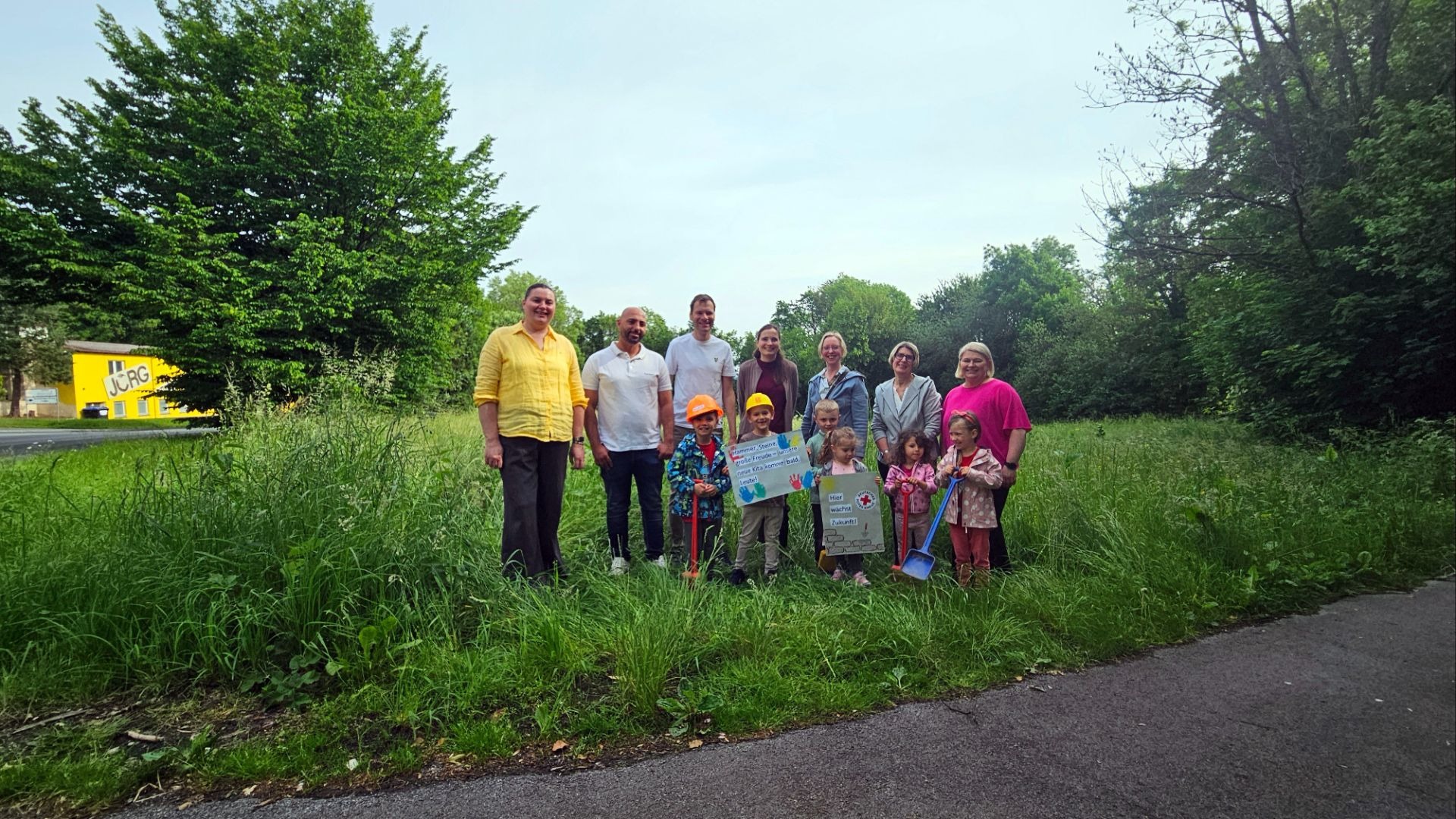 Eine Gruppe Erwachsener und Kinder posiert mit Bauschildern und kleinen Schaufeln auf einer grünen Wiese