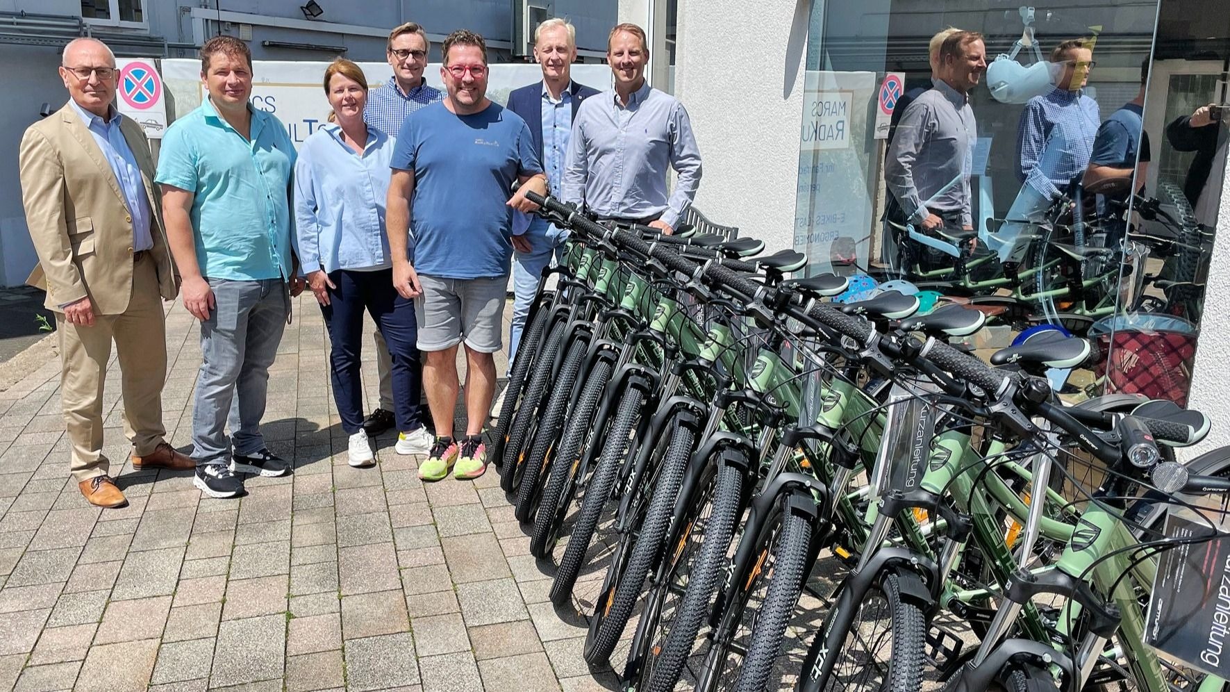 Acht Erwachsene stehen lächelnd neben zehn neuen hellgrünen Mountainbikes auf einem gepflasterten Gehweg