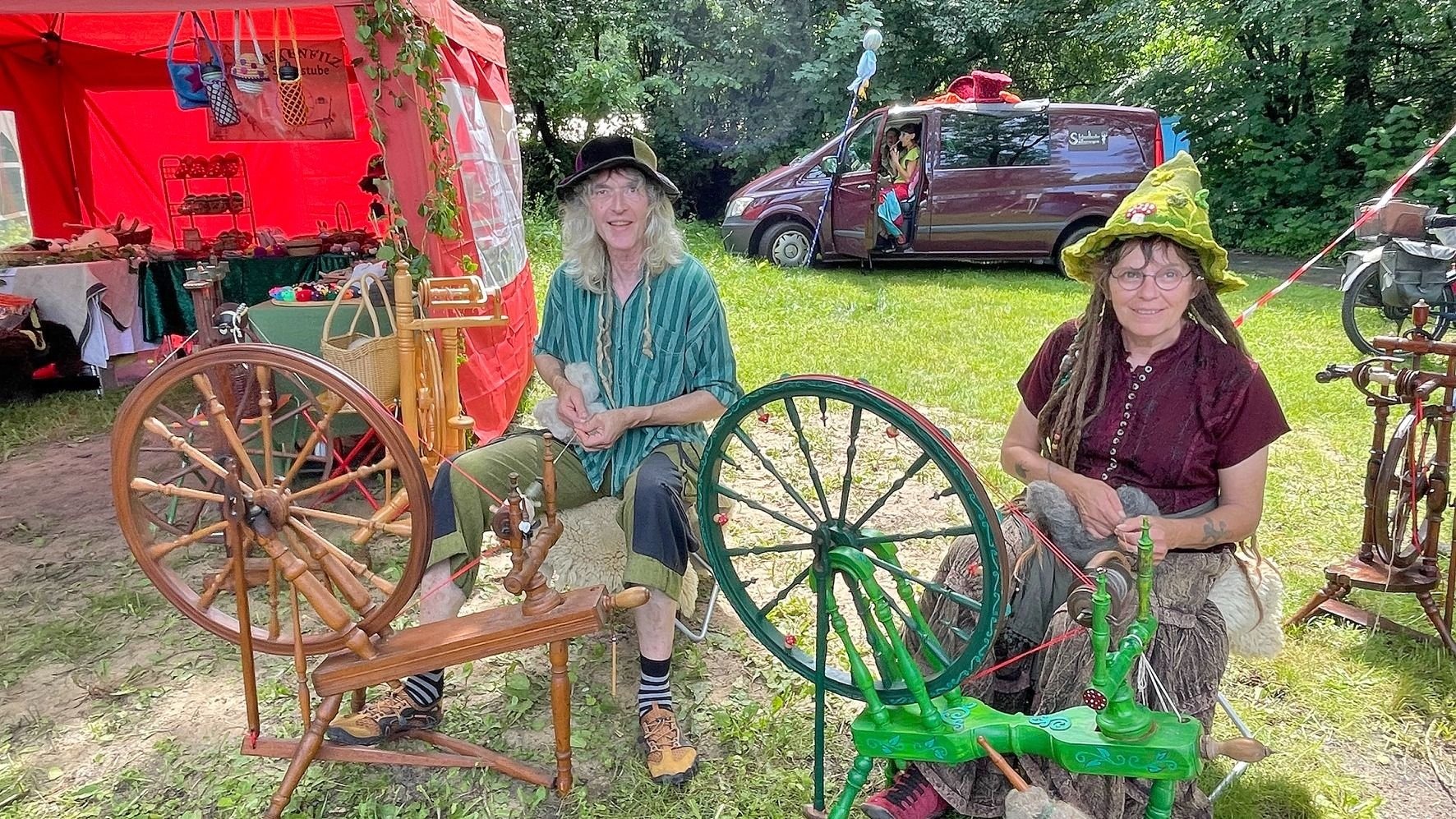 Ein Mann und eine Frau spinnen Wolle an zwei Spinnrädern im Freien vor einem roten Zelt